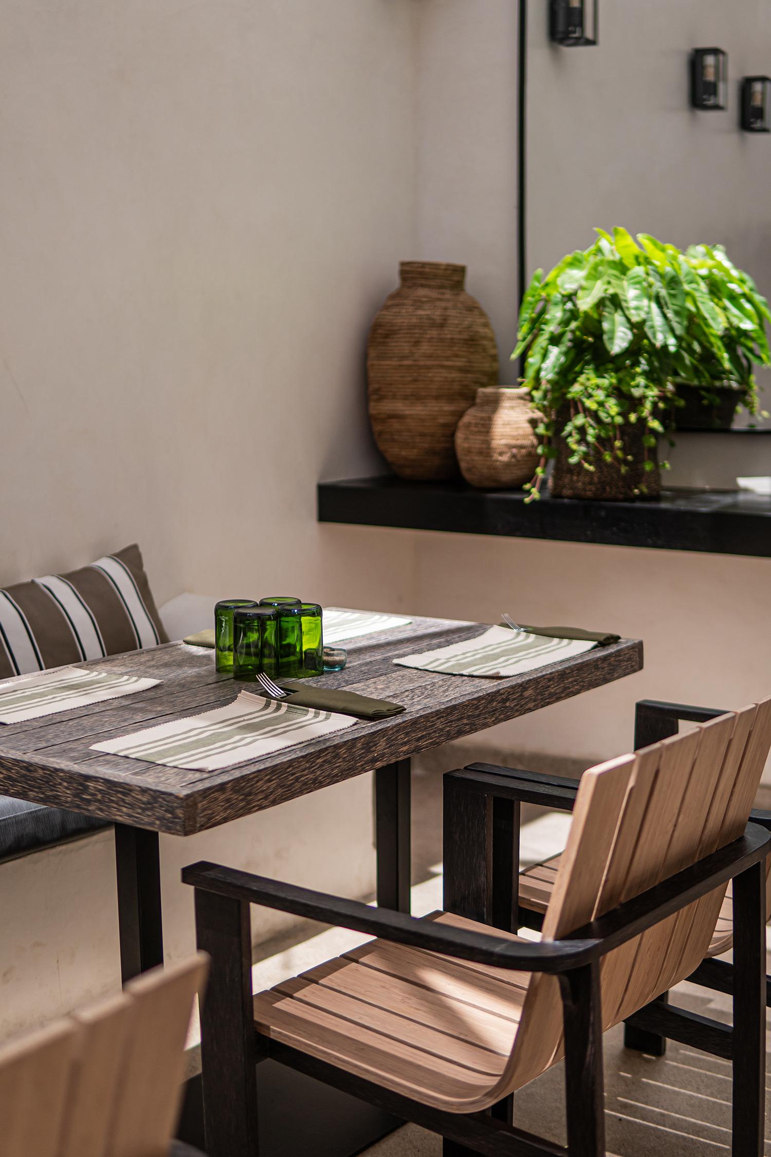 A wooden table is laid with striped placemats, cutlery and green glass tumblers on an outdoor terrace.