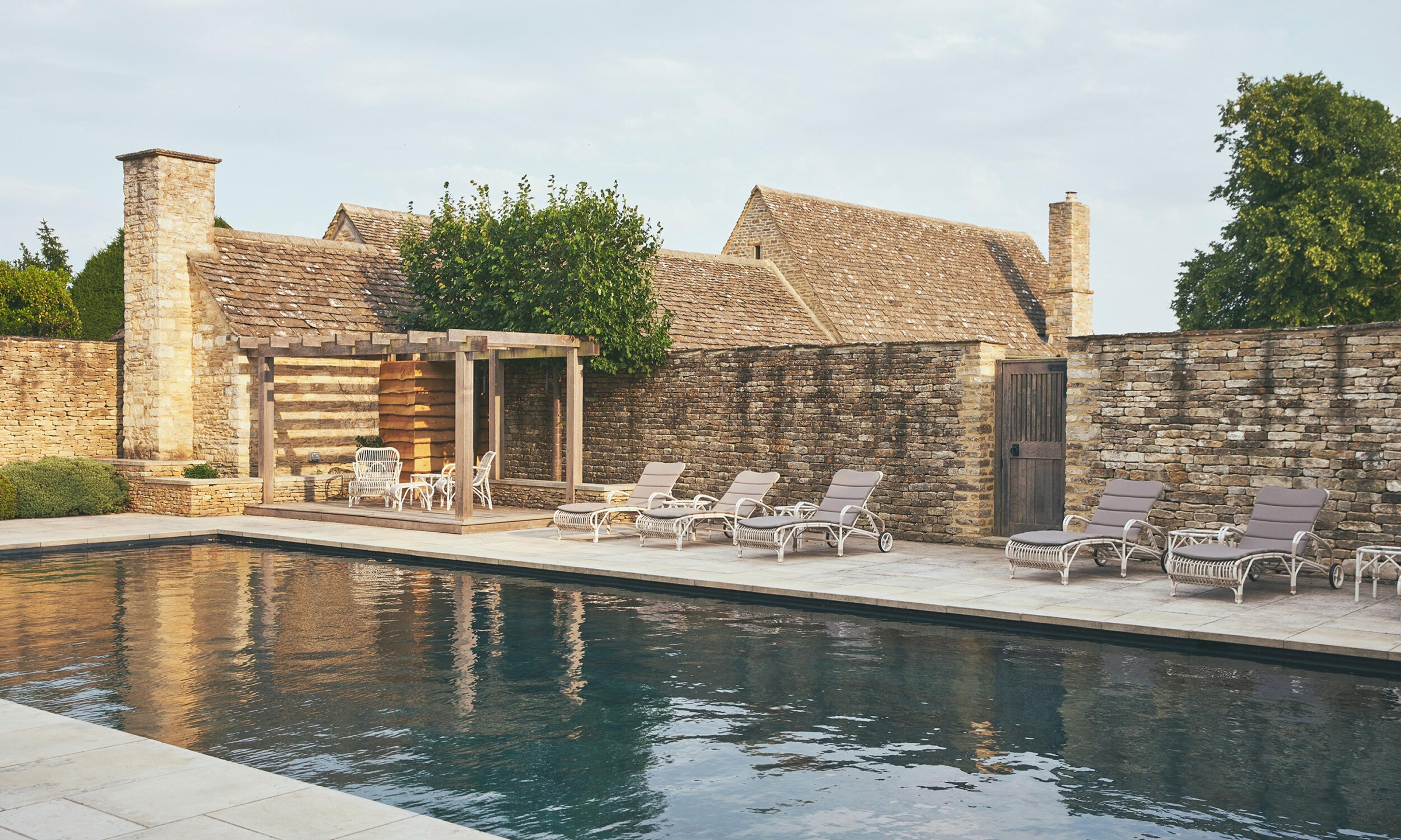 A stone barn and pool house beside a long swimming pool, with sun loungers along the poolside and a wooden pergola at one end, lit by low evening sun.
