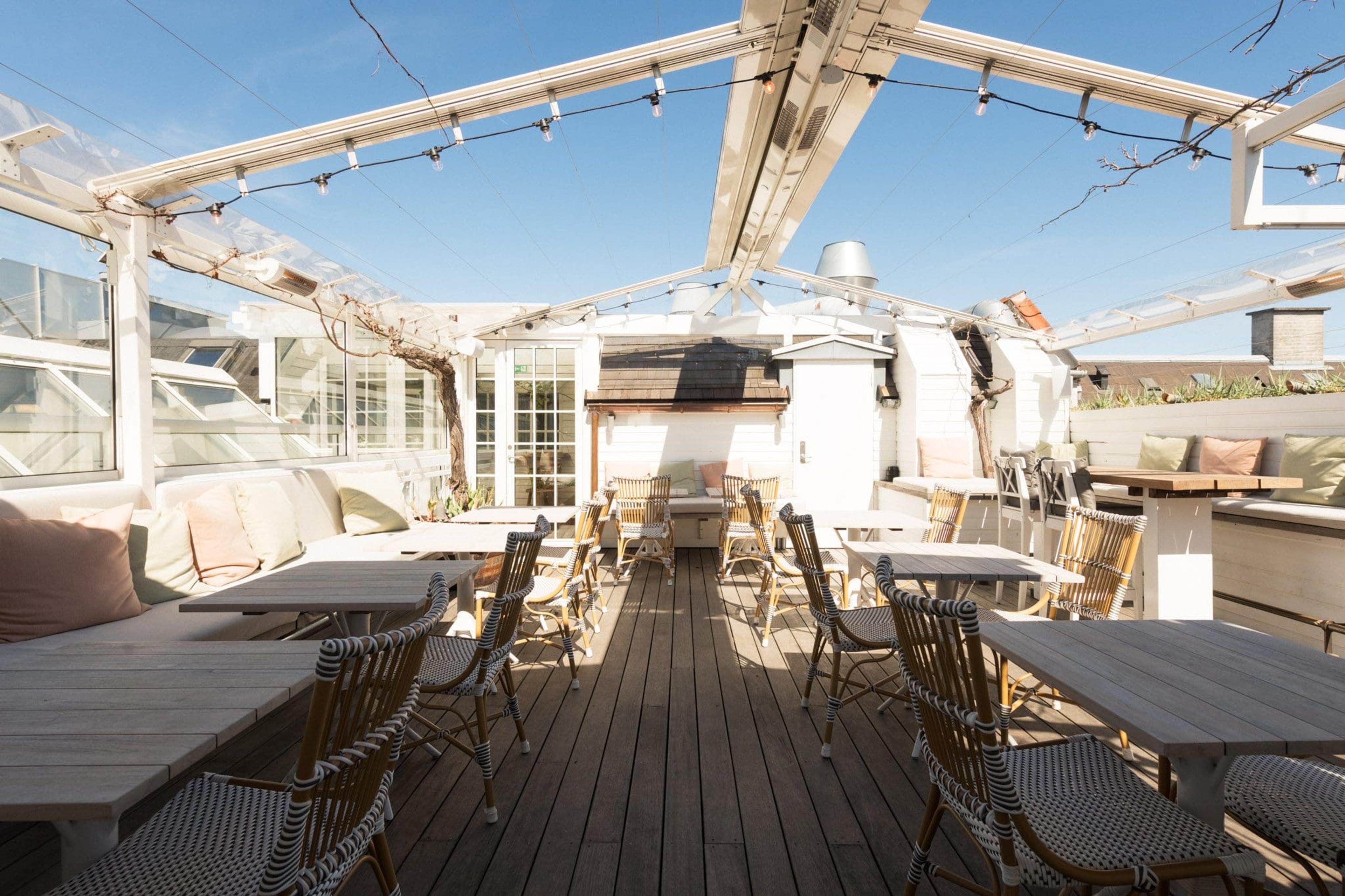 A rooftop terrace with wooden tables and woven chairs under a glass canopy strung with lights, set against a clear blue sky.