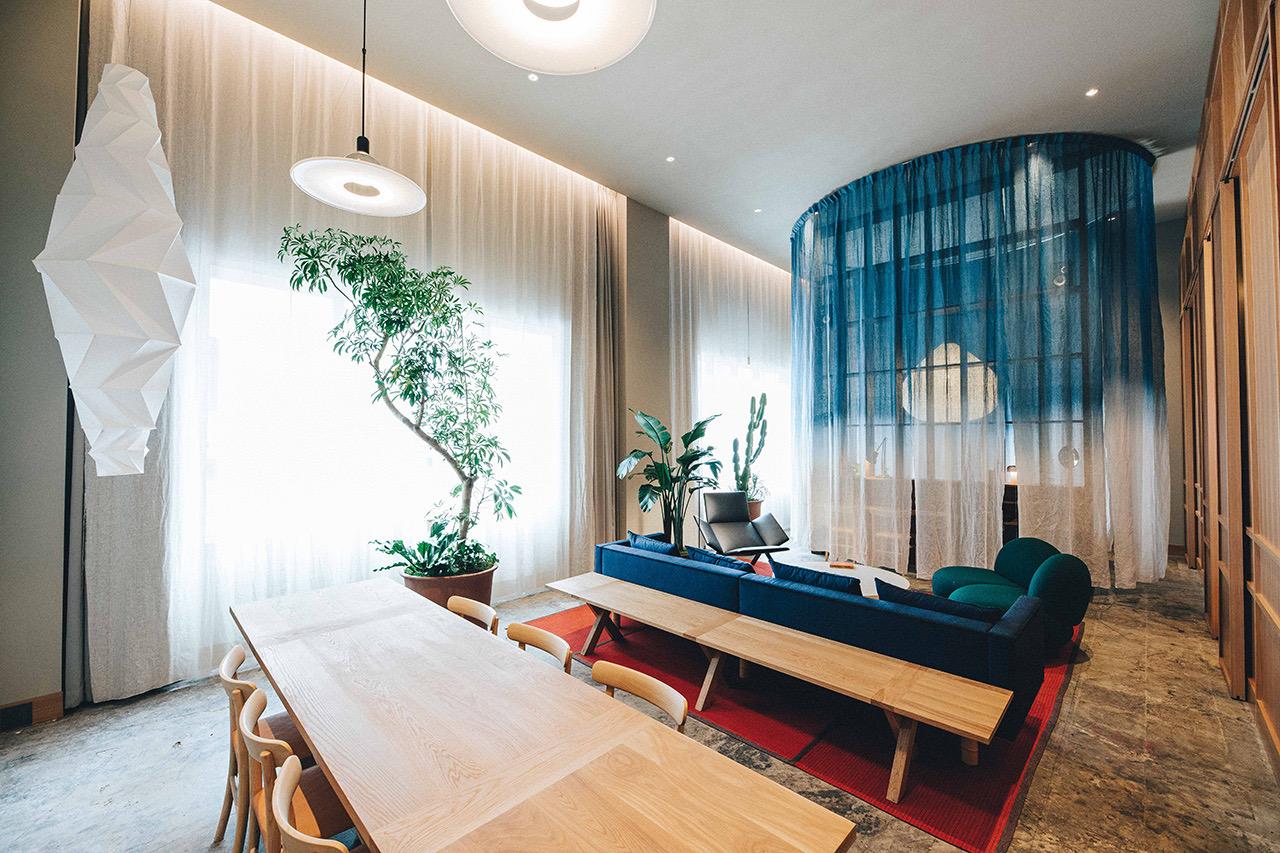 A suite's living area with a long wooden dining table, a navy sofa under a blue dip-dyed curtain enclosure, green armchairs, and potted plants by sheer curtains.