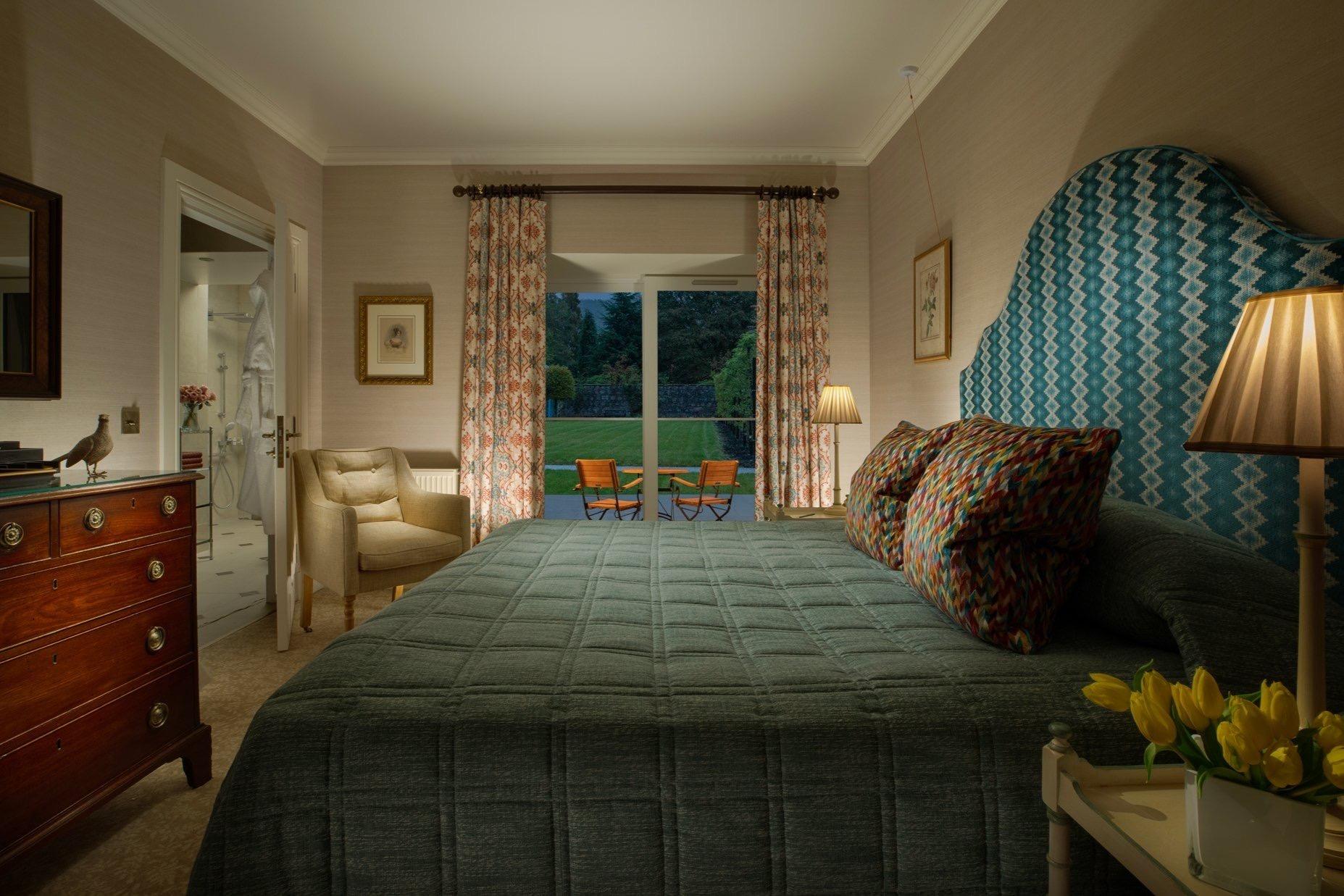 A bedroom with a teal headboard and a vase of yellow tulips, its glass doors open onto a terrace with two chairs and a lawn beyond.
