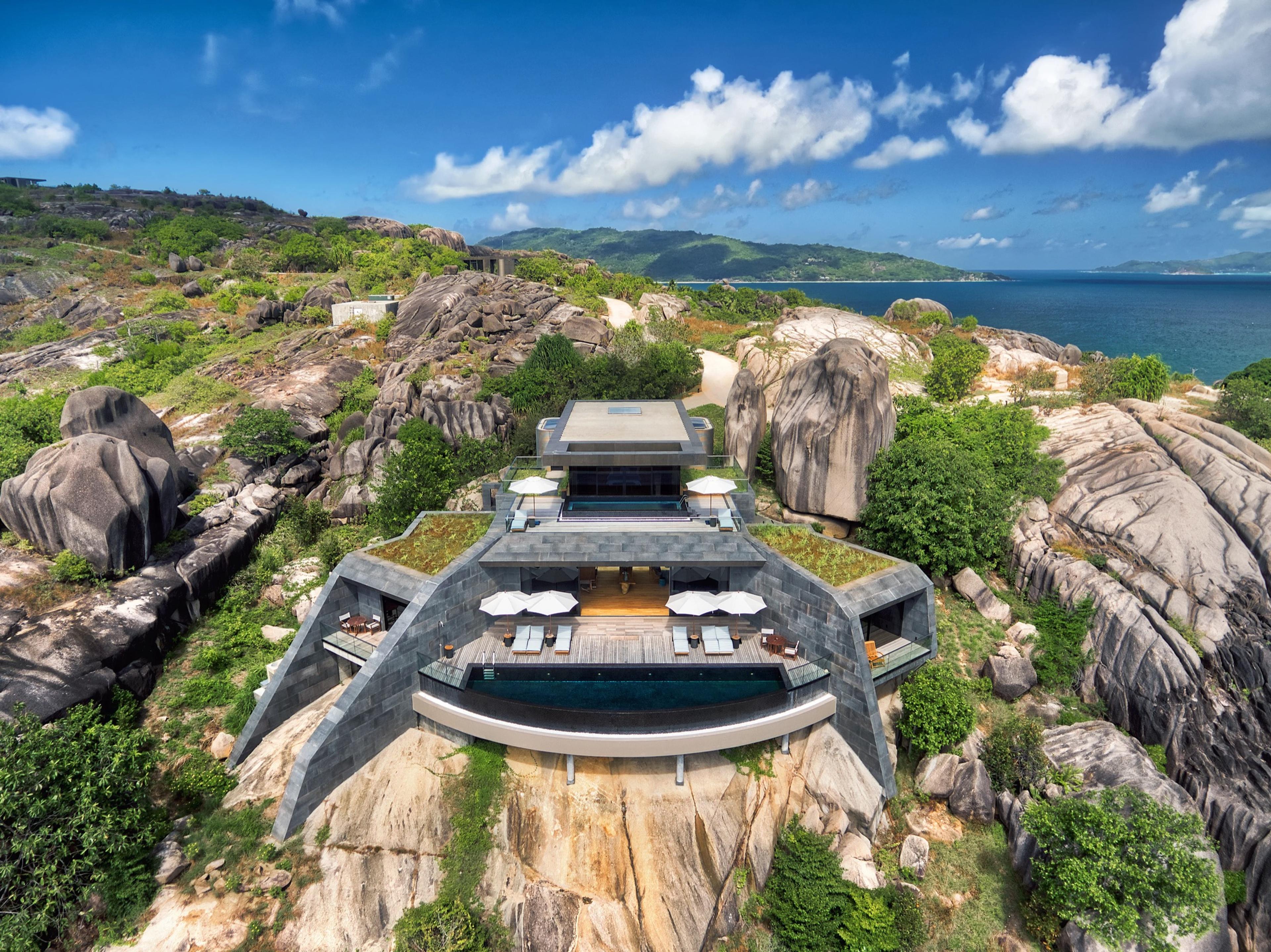 An aerial daytime view of a stone and timber residence built into a granite cliff, with a pool along its lower terrace, boulders and sea on either side under a blue sky.