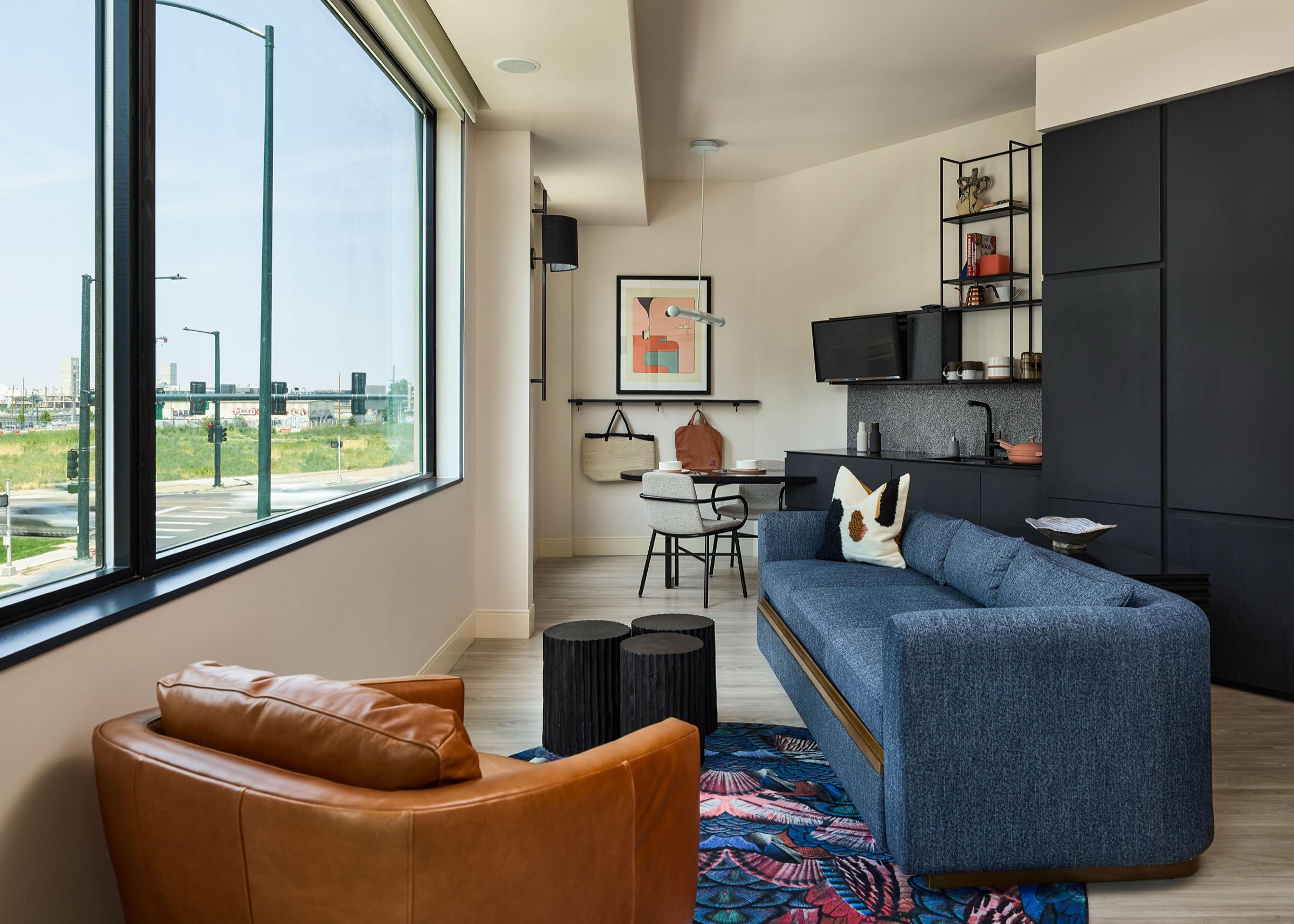 A hotel living room with a brown leather armchair and a navy sofa, floor to ceiling windows looking out over a street and city skyline.