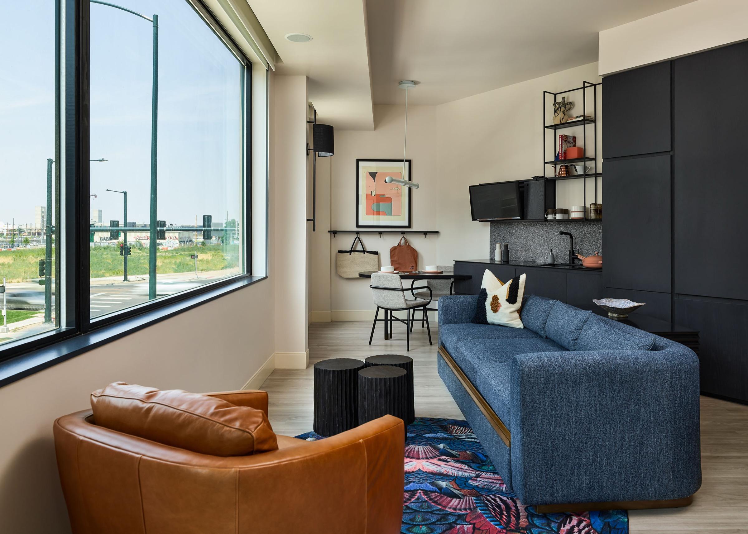 A hotel living room with a brown leather armchair and a navy sofa, floor to ceiling windows looking out over a street and city skyline.