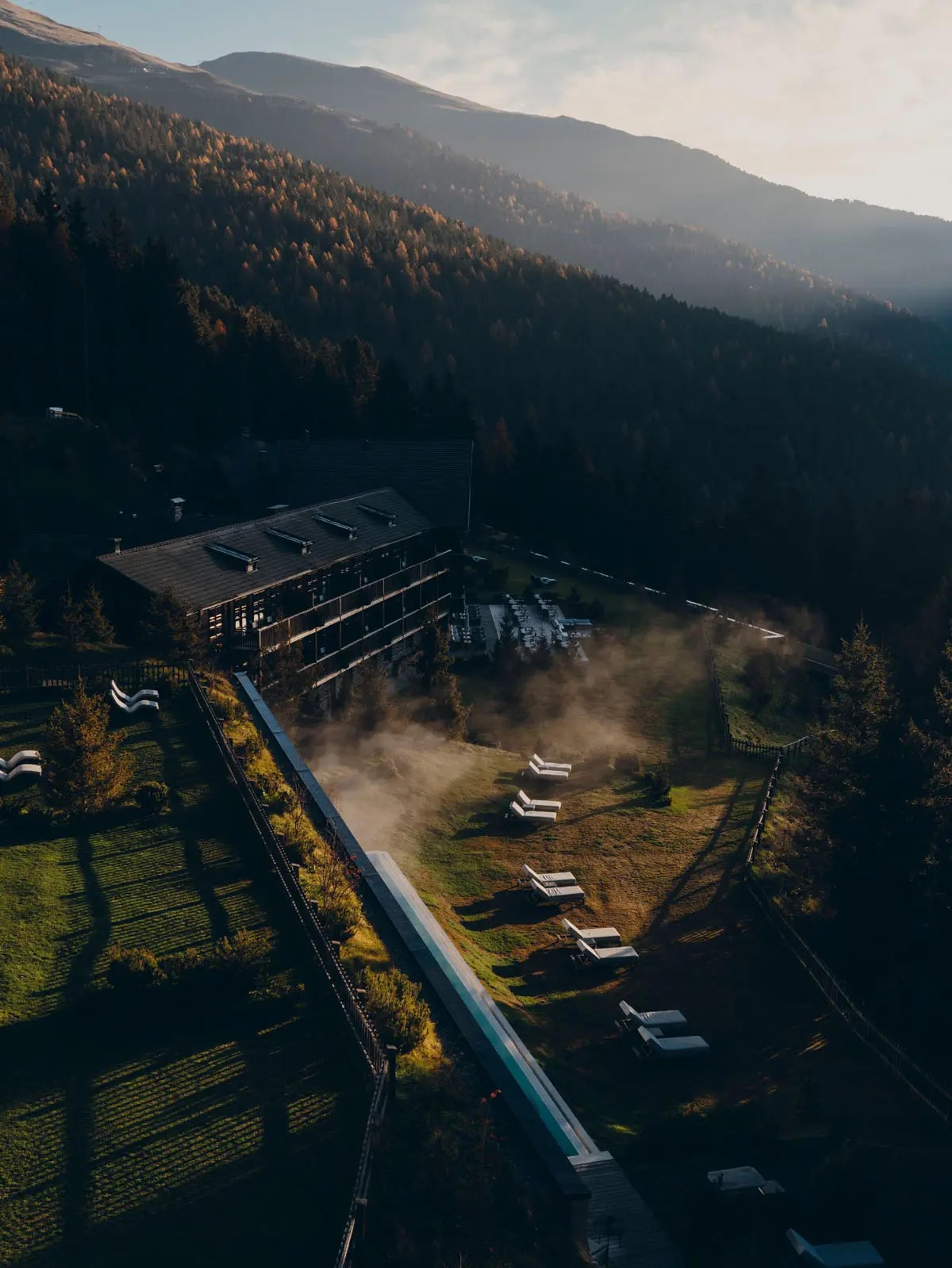 An aerial view at dusk shows the hotel building beside a long pool, mist rising over the grounds against forested mountains.