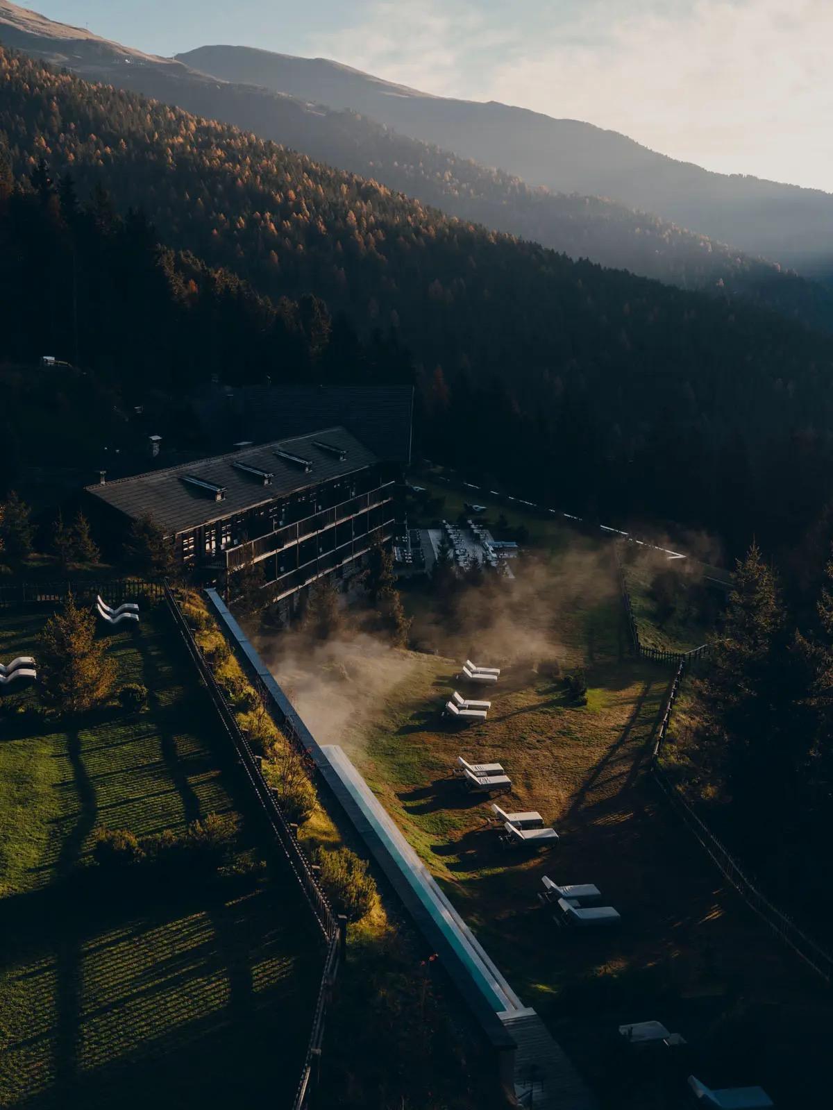 An aerial view at dusk shows the hotel building beside a long pool, mist rising over the grounds against forested mountains.