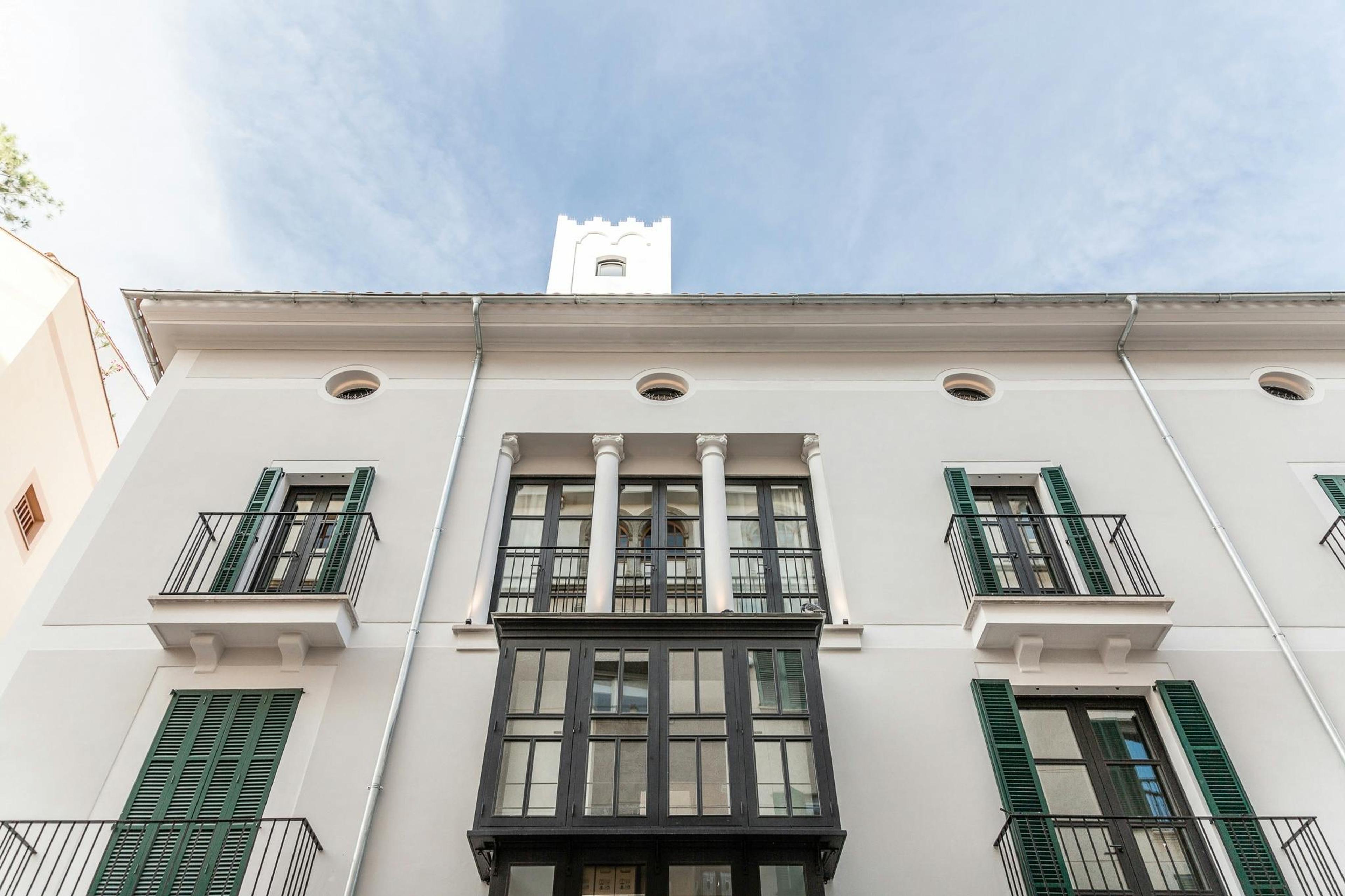 The cream stone facade of the hotel seen from below, with green-shuttered windows, wrought-iron balconies and a crenellated tower against the sky.
