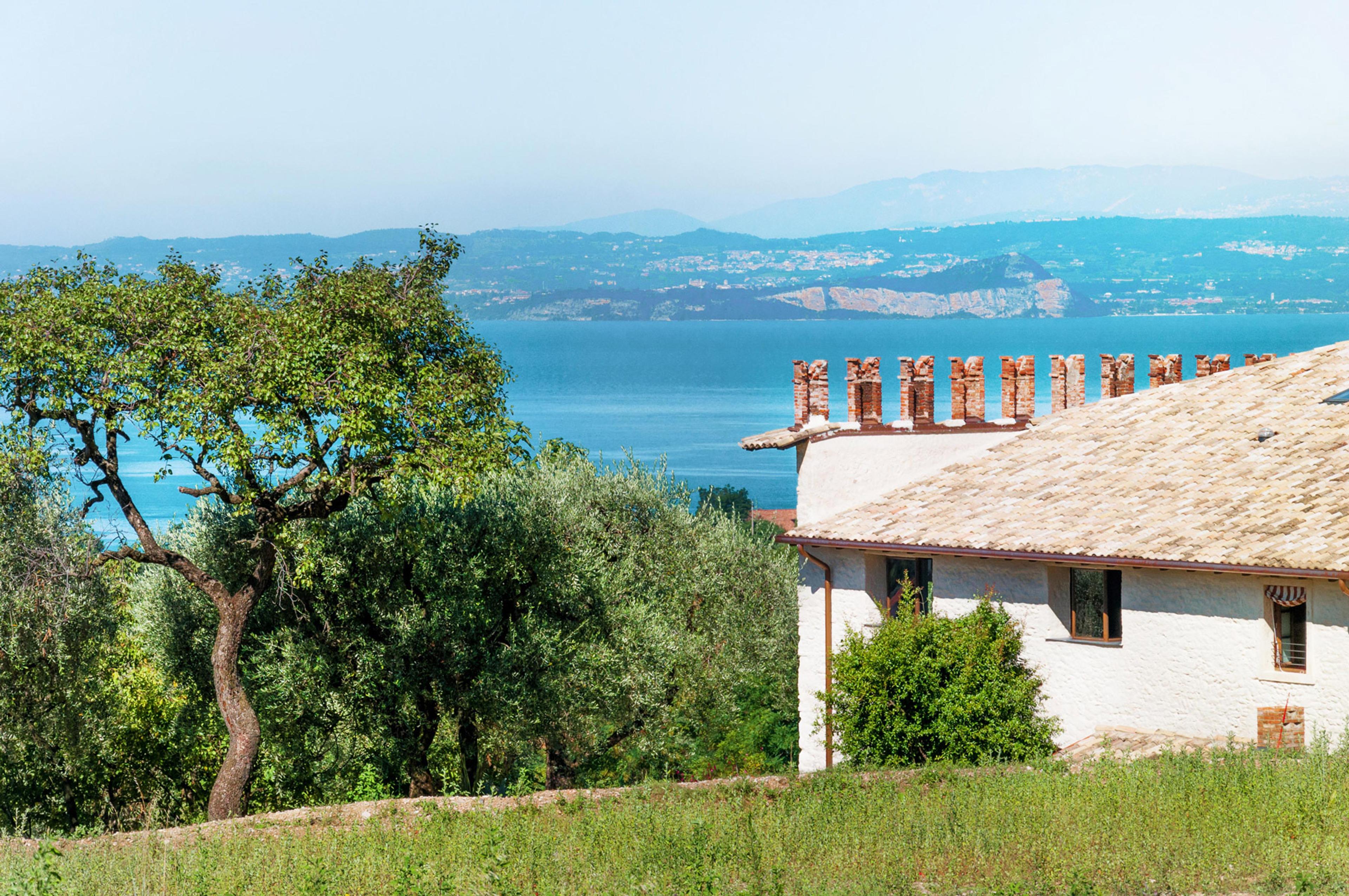 The white stuccoed corner of a tiled-roof building with a row of brick crenellations along the ridge, an olive tree in front and a lake with mountains beyond.