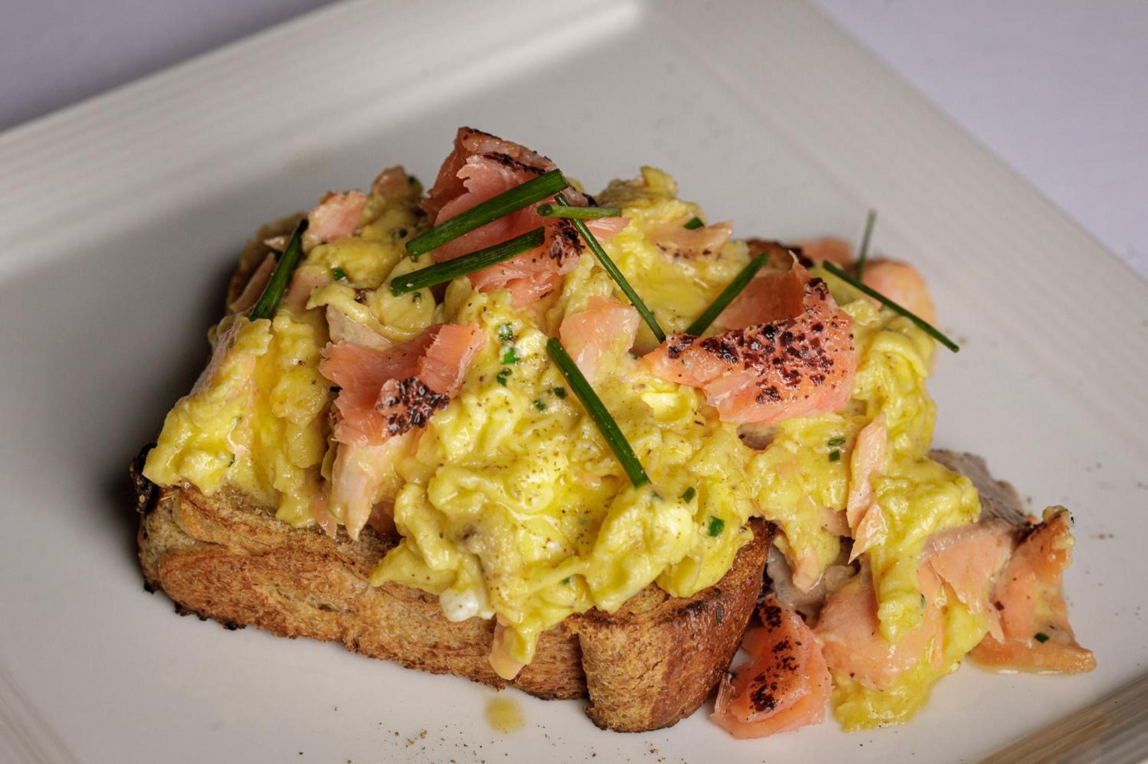 A slice of toast topped with scrambled egg, smoked salmon and chopped chives, on a white plate.