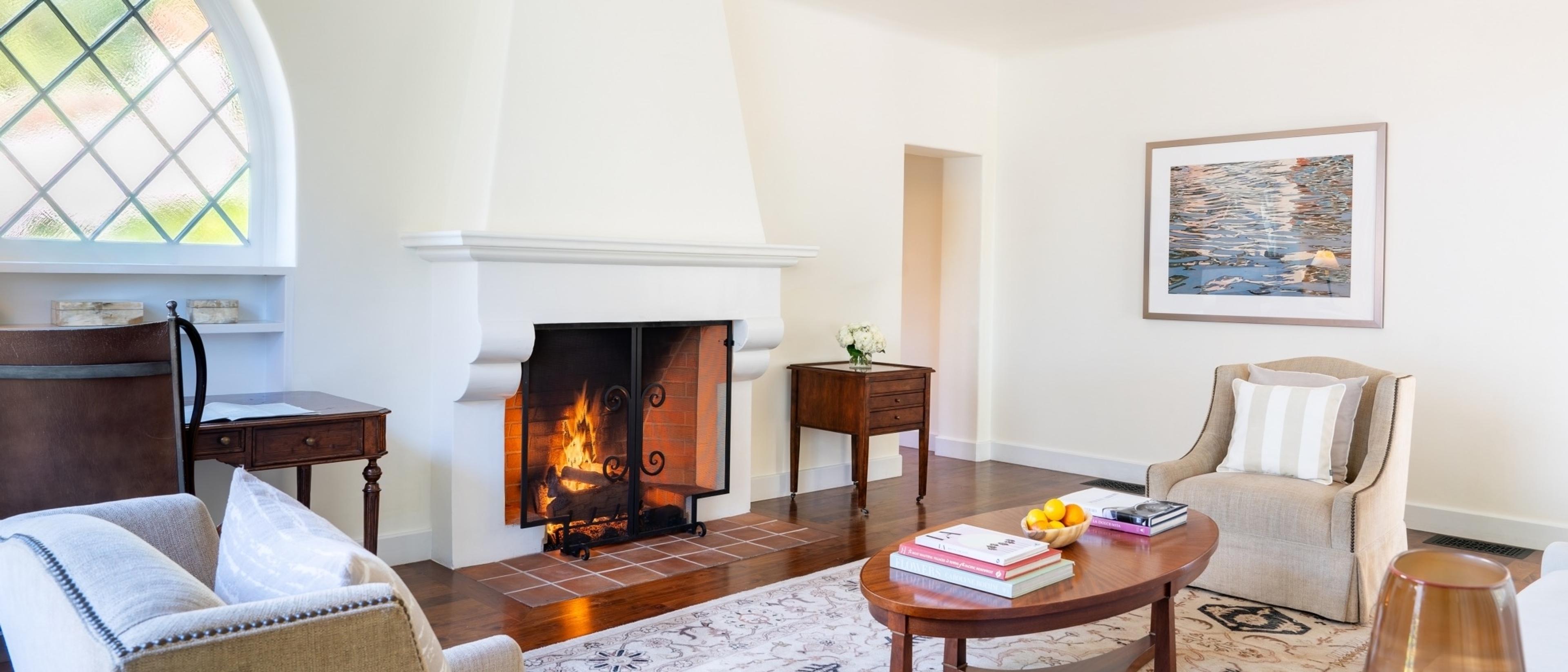 A sitting room with a lit fireplace, two upholstered armchairs, a wooden coffee table holding books and a bowl of fruit, and an arched leaded-glass window.