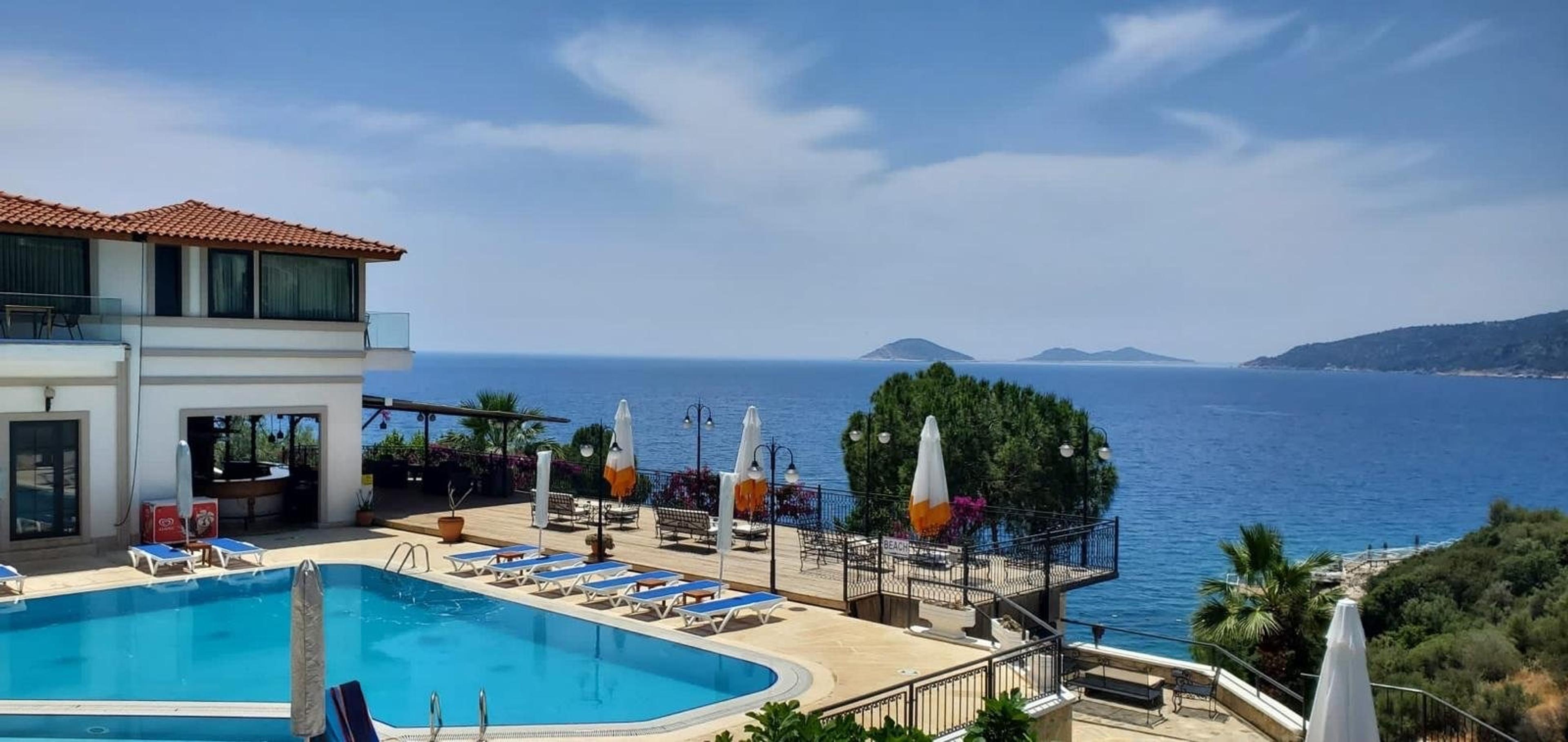 An aerial view of a hotel's restaurant building, pool, and car park on a cliff edge, with wooden sunbathing platforms over the sea beyond.
