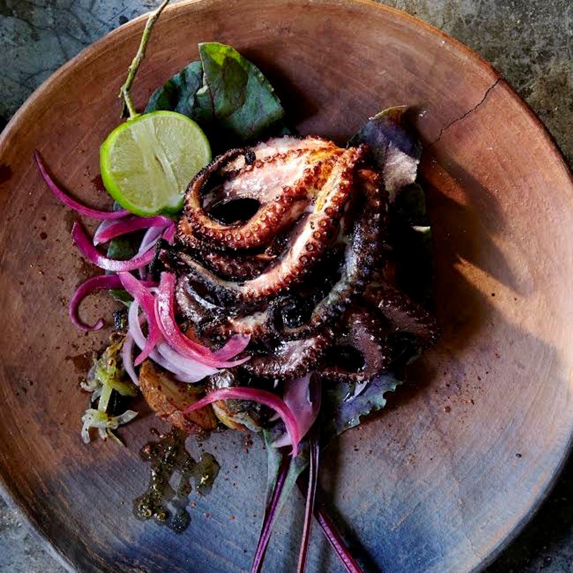 Charred octopus with pickled red onion and a lime half on a wooden plate