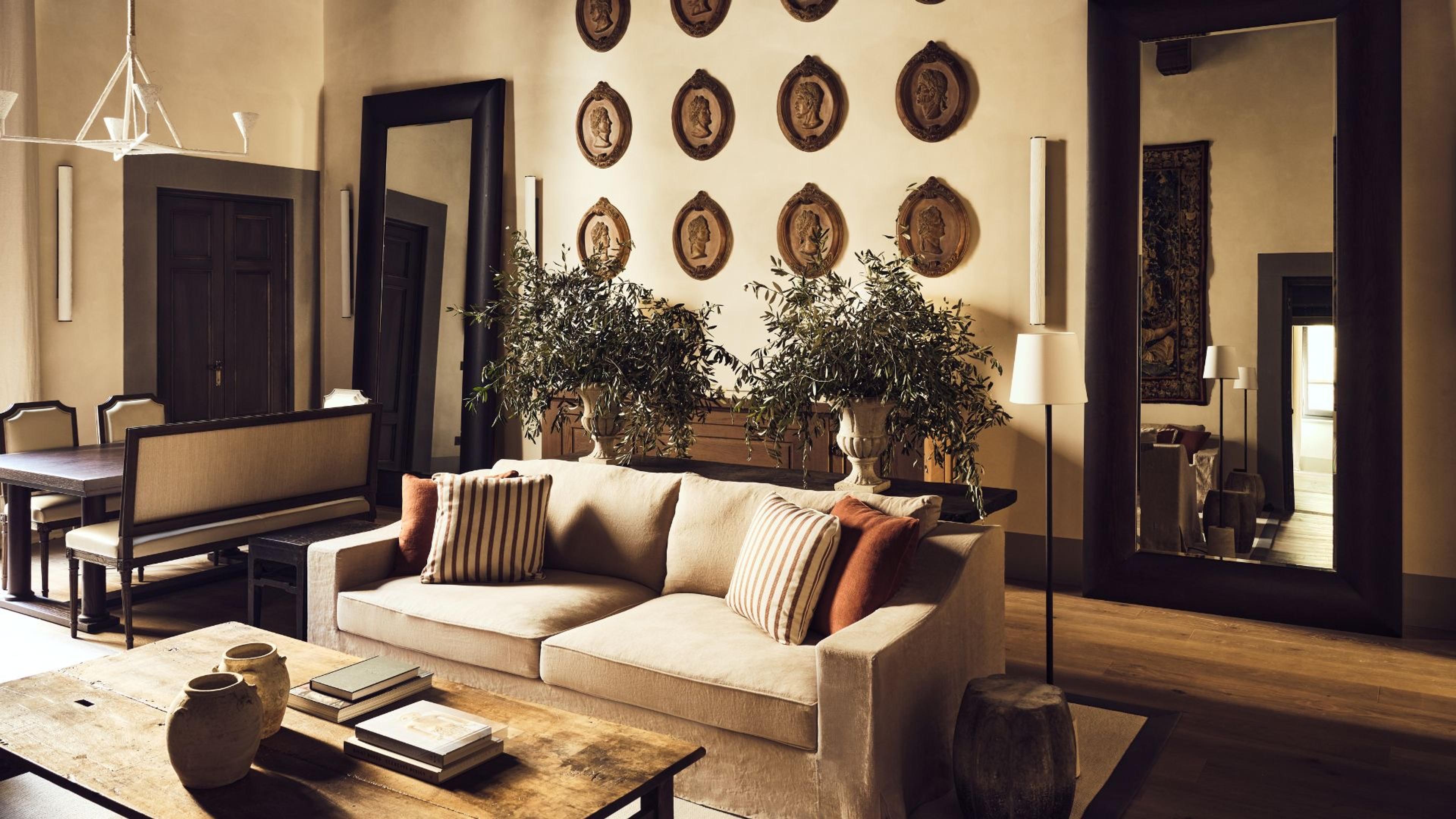 A cream sofa with striped and rust cushions in a sitting room, facing a wall hung with eight oval bronze relief medallions, beside a dining table and a large mirror.