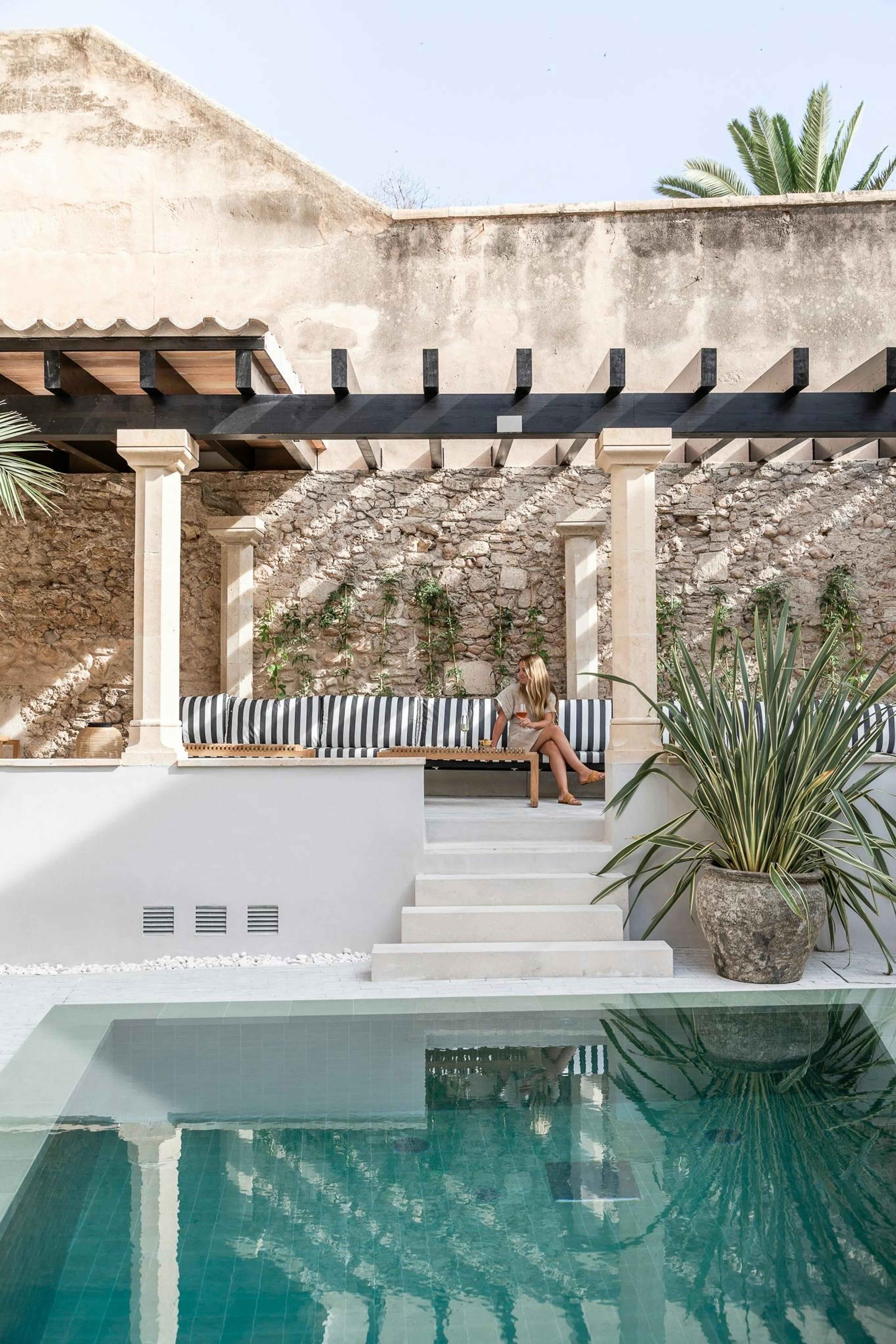 A plunge pool in a stone-walled courtyard, with a shaded pergola seating area and a woman sitting at a distance beyond the pool.