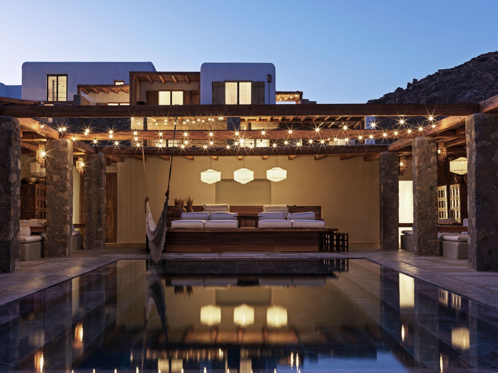 At dusk, a two-storey whitewashed building glows from within behind a long pergola strung with lights, a hammock hanging over a dark reflecting pool in front of a built-in lounge.