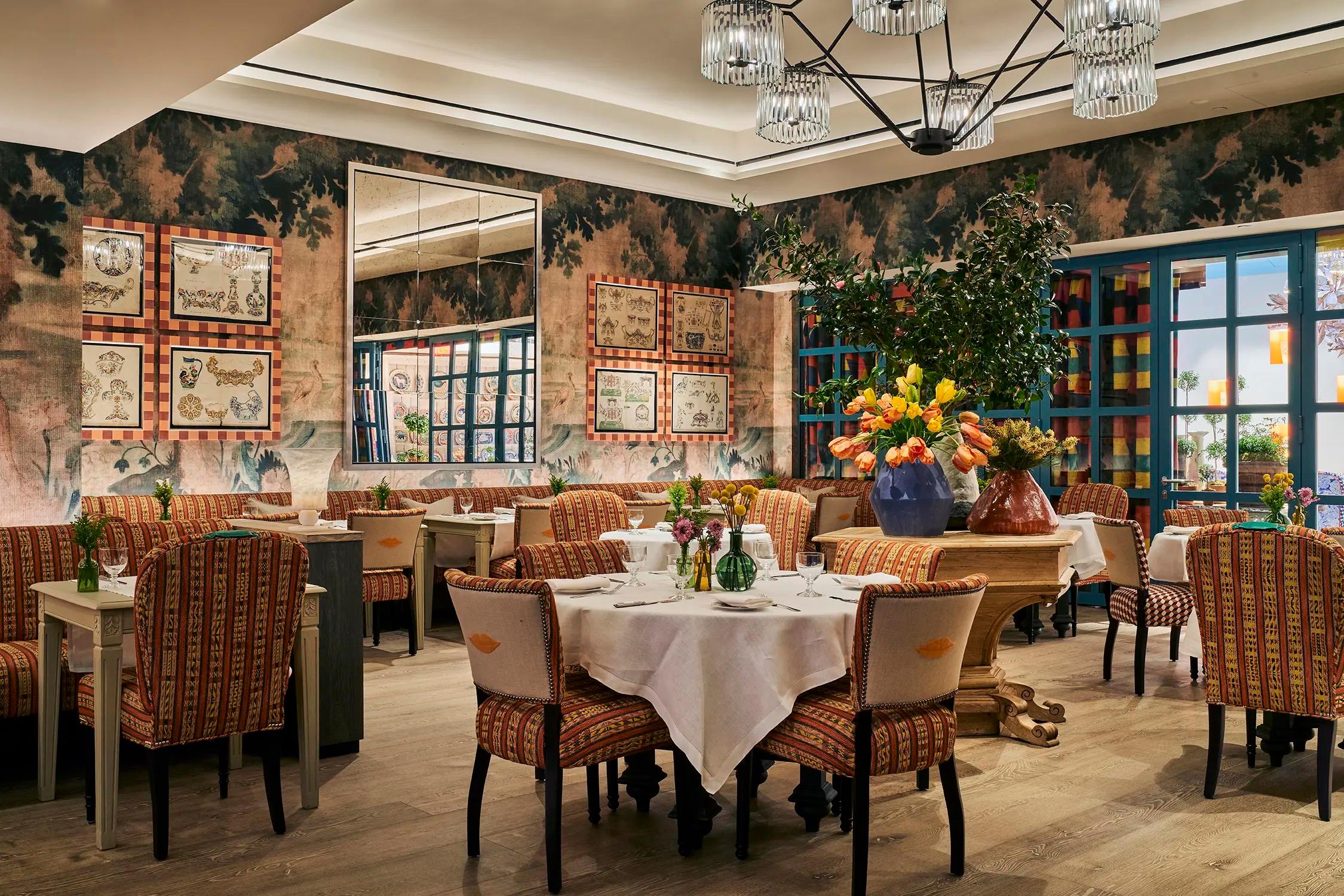 A restaurant dining room with patterned upholstered chairs around tables laid in white linen, under a mural-papered wall and a crystal chandelier.