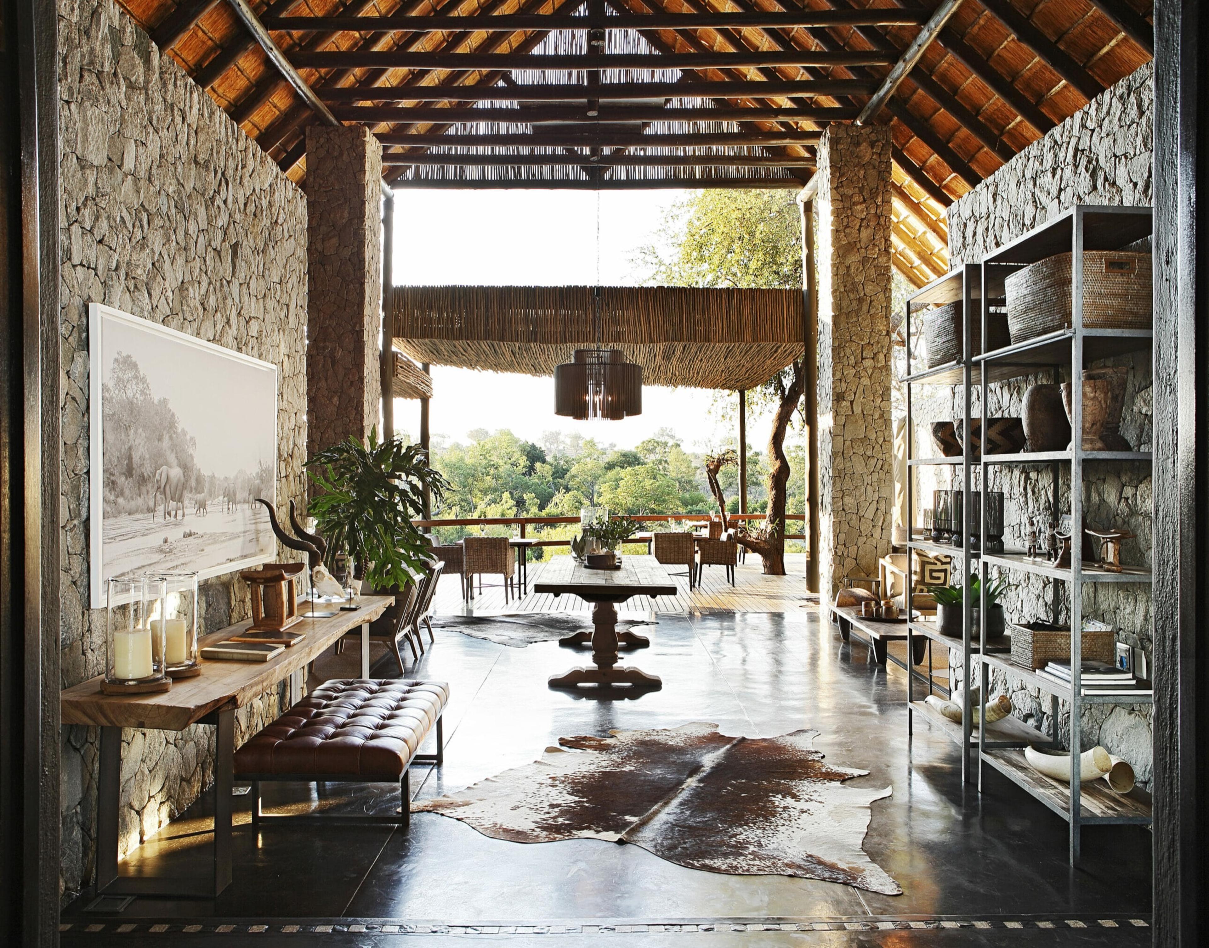 a stone-walled lodge entrance with a long wooden table, a leather bench and a bowl of candles, opening onto a thatched deck overlooking a canopy of trees