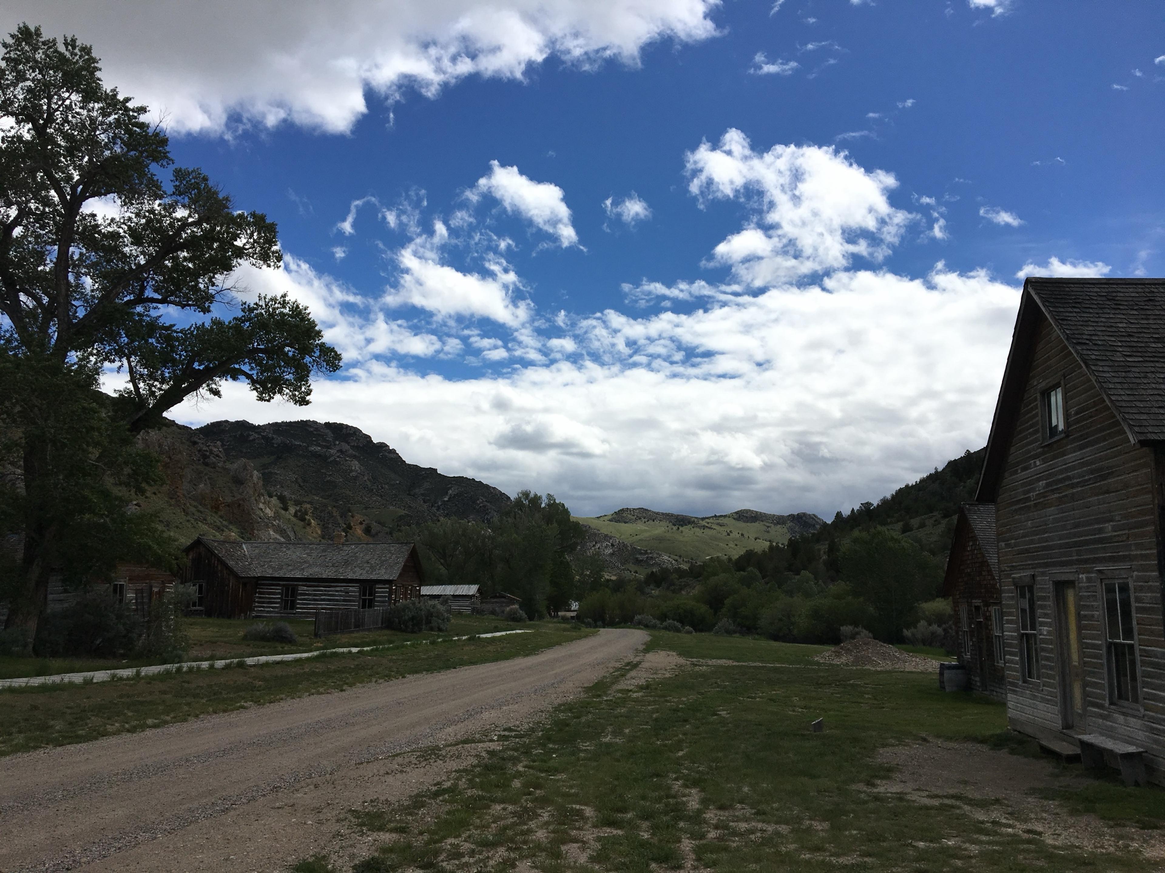 Weathered wooden buildings beside a dirt road, with green hills behind and big white clouds