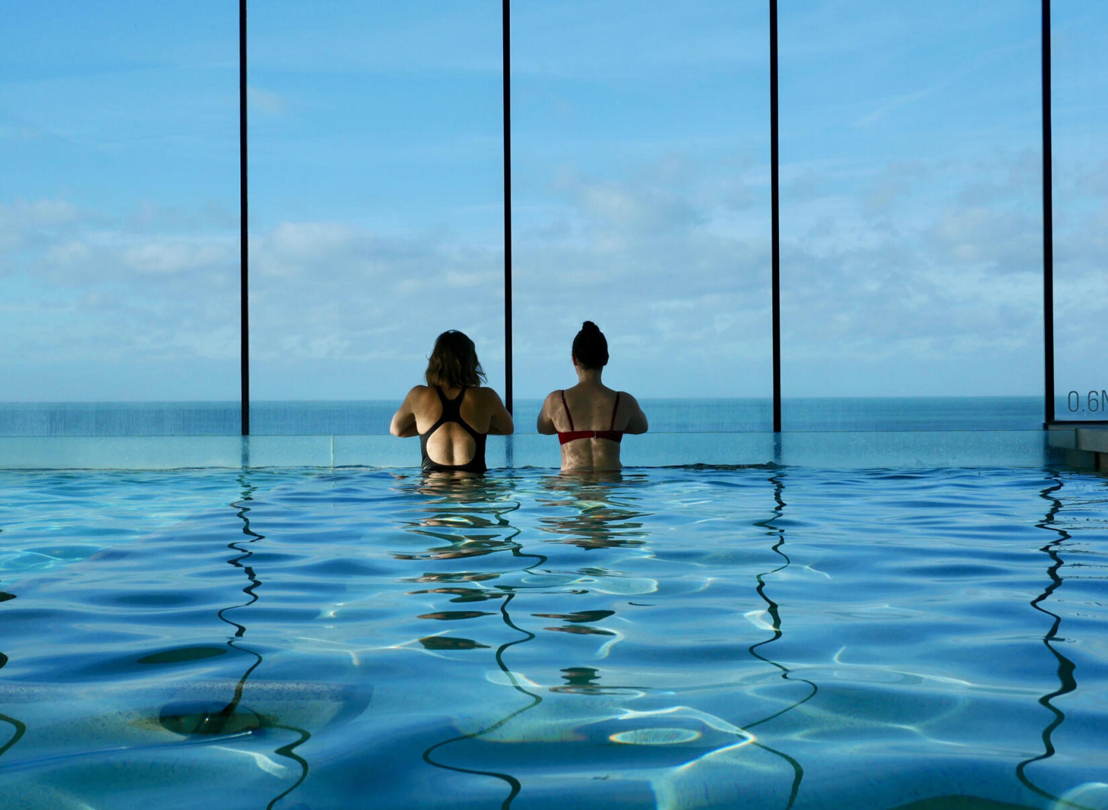 Two women standing in an infinity pool with their backs to the camera, looking out at the sea.