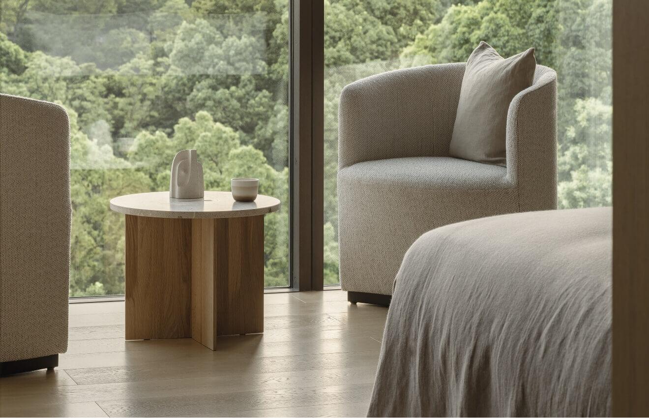 a hotel room sitting area with two armchairs and a round wood side table beside a window looking onto dense treetops, with the corner of a grey-covered bed in the foreground