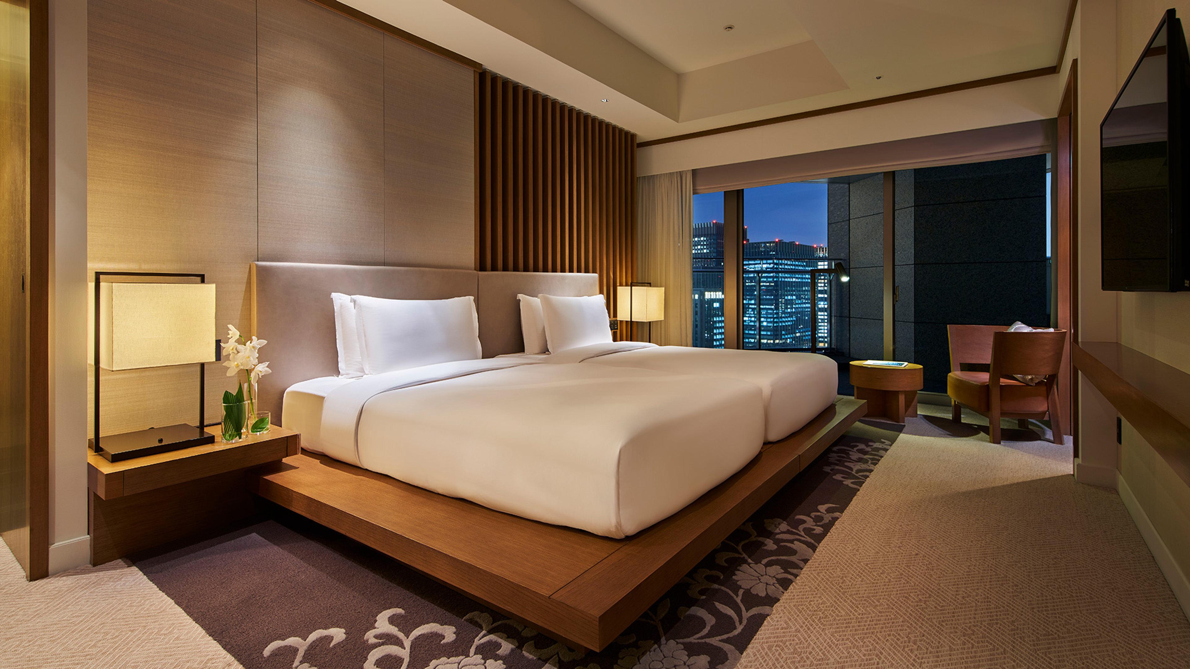 A hotel bedroom at night, a wide bed between two lit lamps and floor to ceiling windows showing the city skyline.
