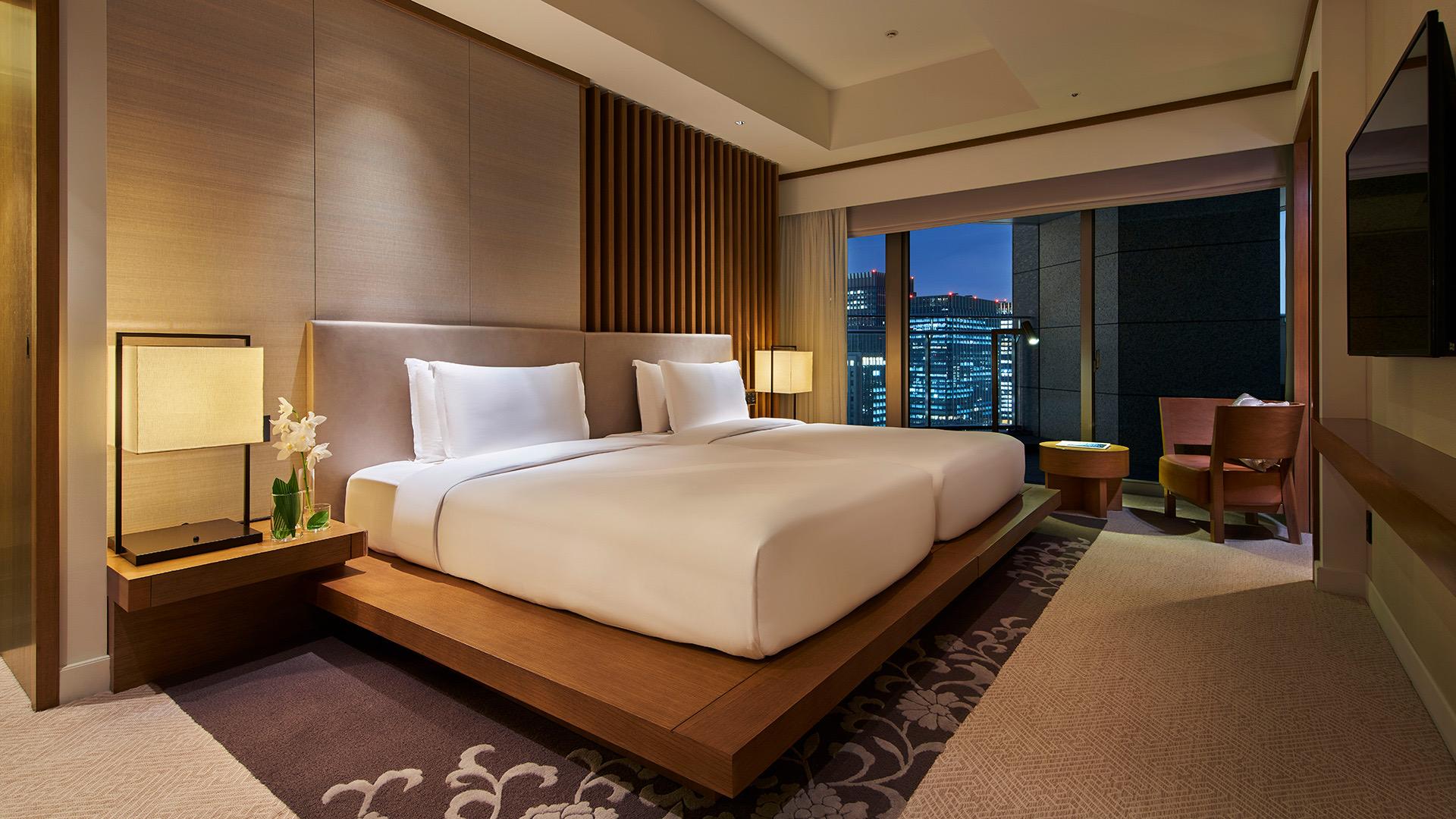 A hotel bedroom at night, a wide bed between two lit lamps and floor to ceiling windows showing the city skyline.