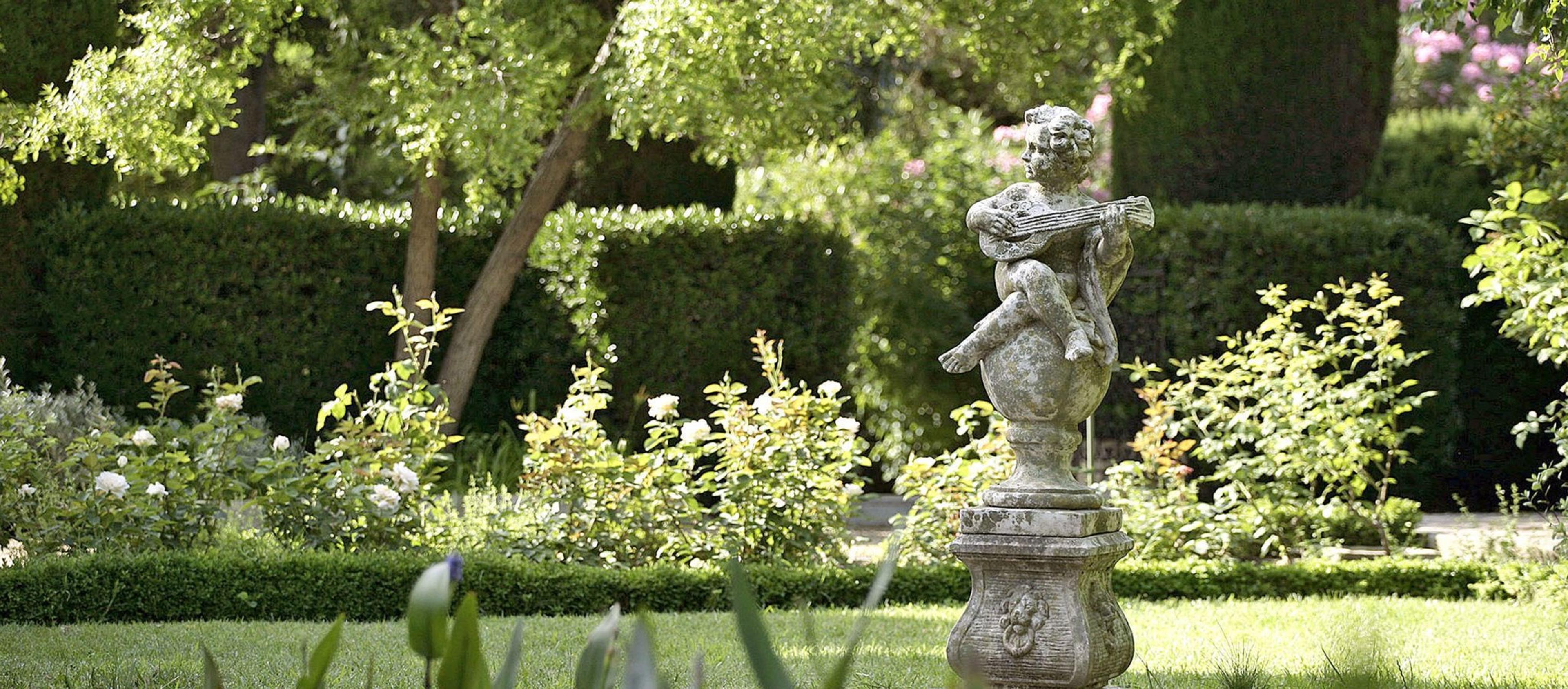 A stone cherub statue playing a lute stands among roses and clipped hedges in a sunlit garden.