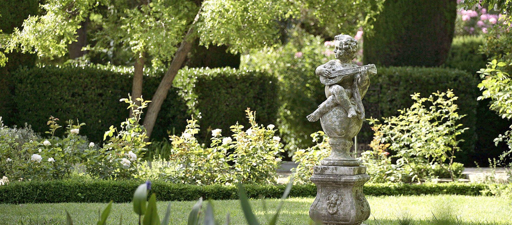 A stone cherub statue playing a lute stands among roses and clipped hedges in a sunlit garden.