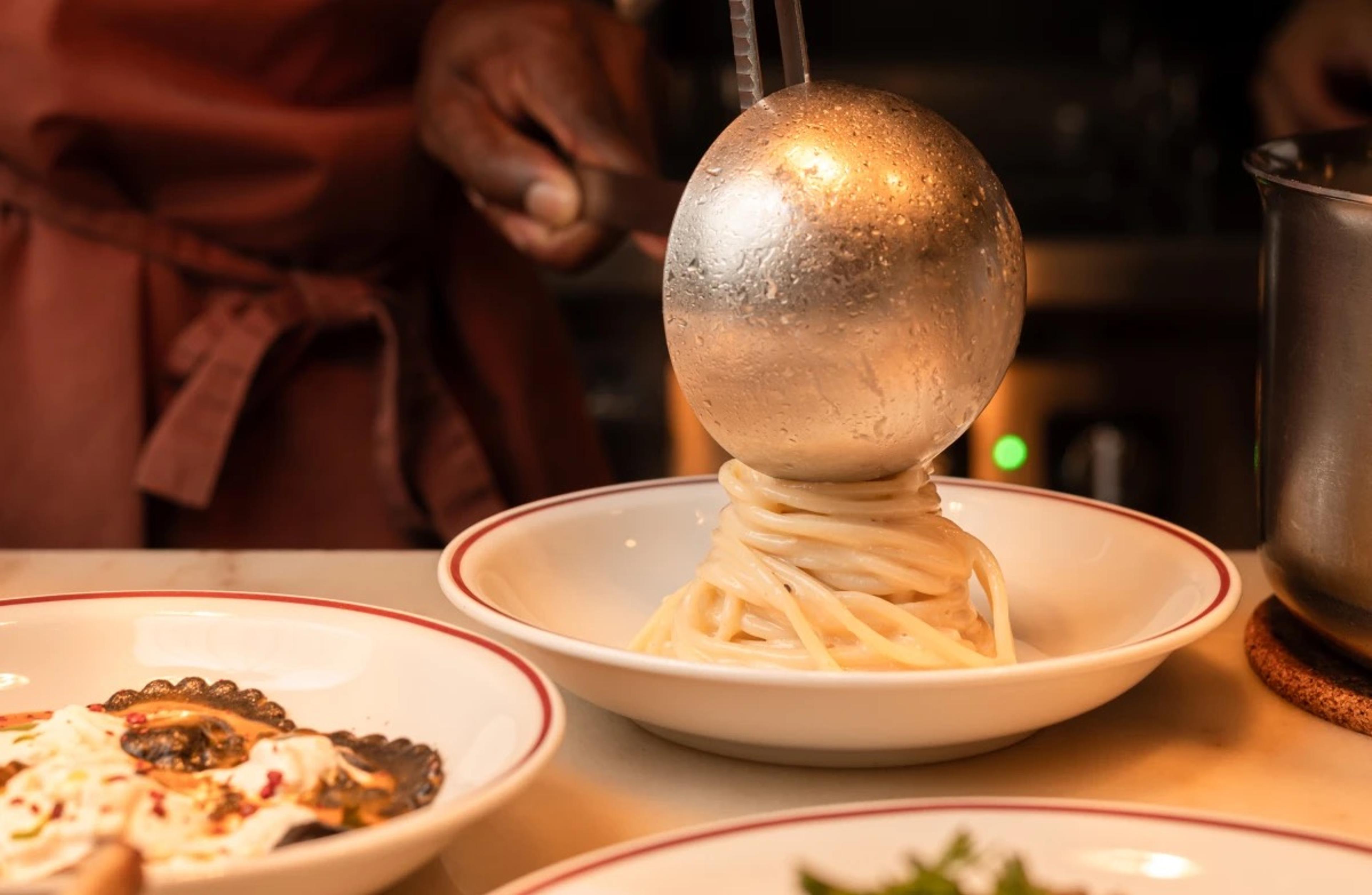 A silver ladle lifts a swirl of spaghetti onto a white bowl with a red rim, with a cook's hand and apron in the background and another plate in the foreground.