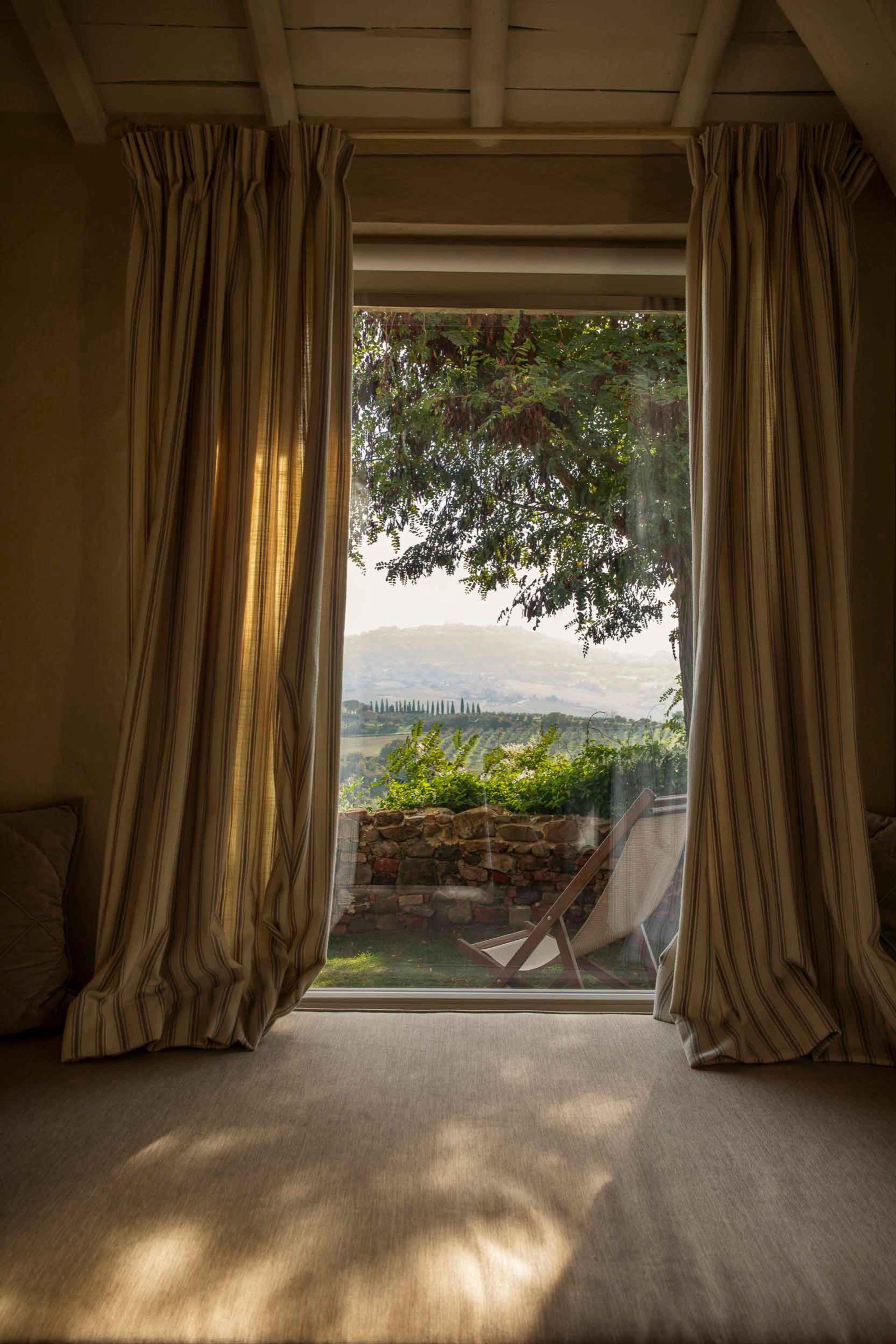 A bedroom with striped curtains framing an open door onto a garden, a tree and hazy hills beyond, with a deck chair outside.