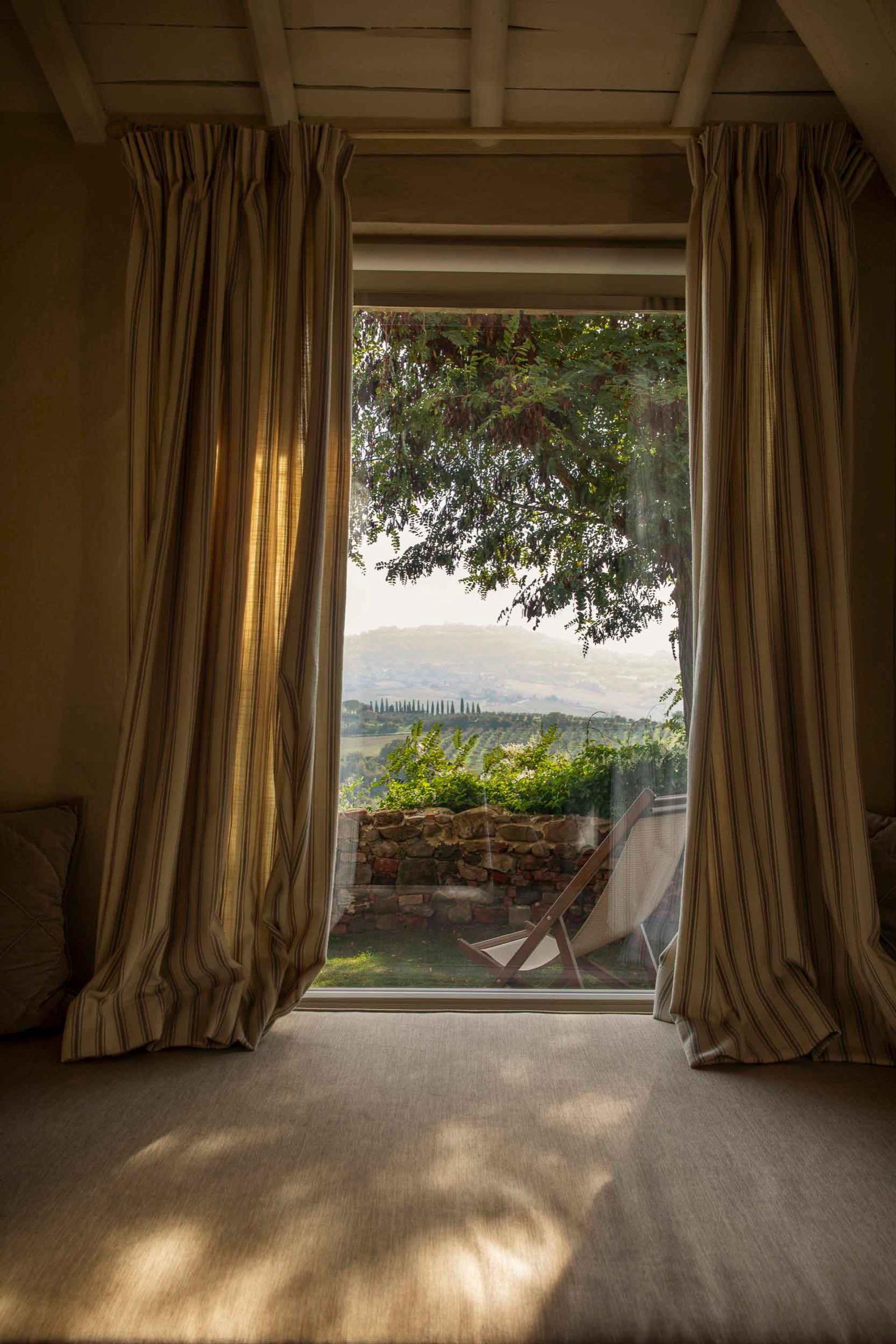 A bedroom with striped curtains framing an open door onto a garden, a tree and hazy hills beyond, with a deck chair outside.