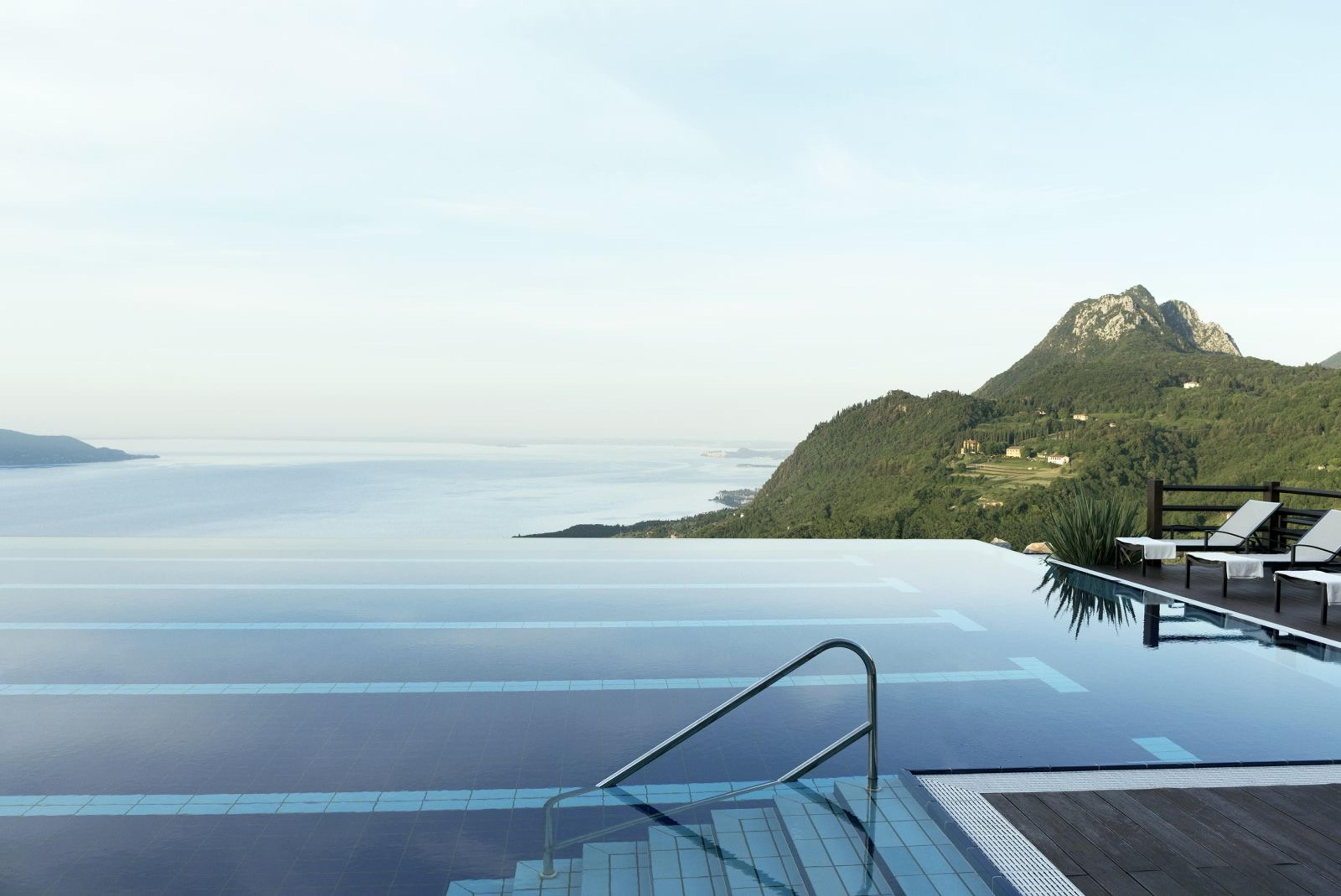 An empty infinity pool stretching towards the lake and a line of wooded mountains, with two loungers set on a deck to the side.