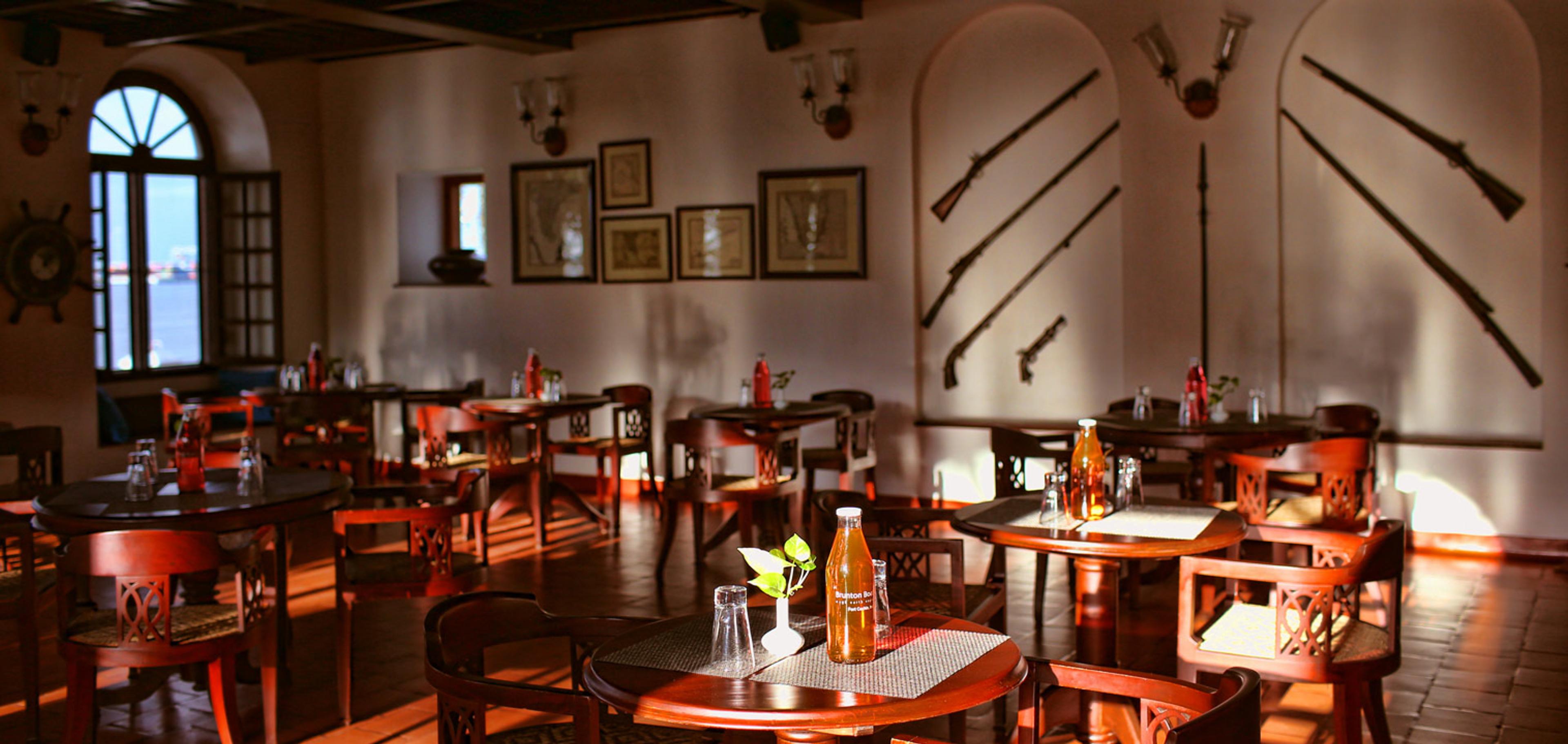 An empty dining room with dark wood tables and chairs on a red tiled floor, arched windows, and antique rifles and a ship's wheel mounted on the wall.