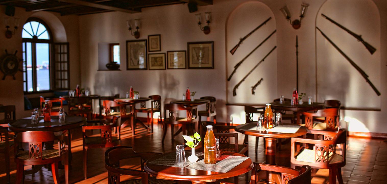 An empty dining room with dark wood tables and chairs on a red tiled floor, arched windows, and antique rifles and a ship's wheel mounted on the wall.