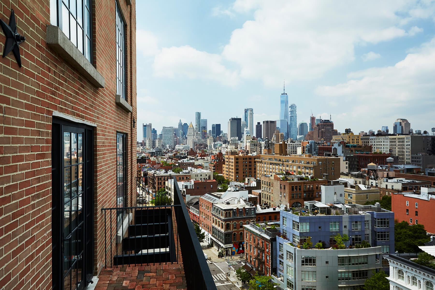 A view from a rooftop terrace over Manhattan rooftops to the World Trade Center, with a brick wall and iron railing in the foreground.