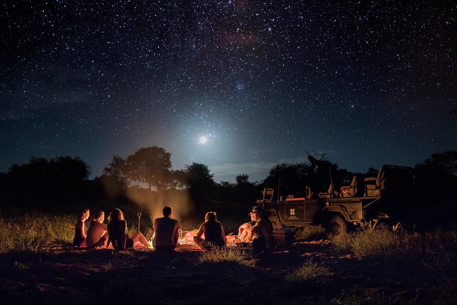 a group of people seen from behind sitting around a campfire at night under a bright starry sky, with a jeep parked alongside