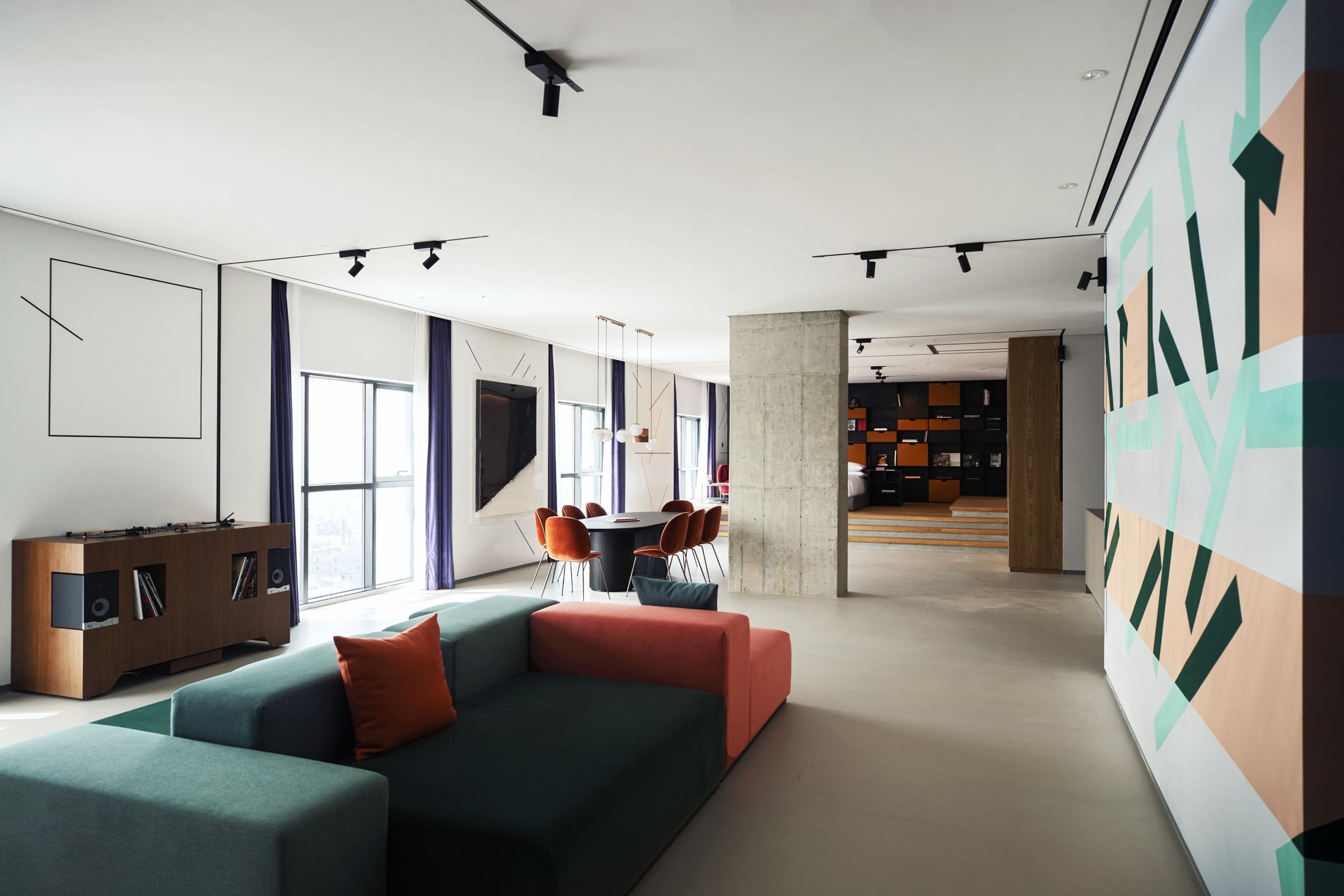 A hotel suite lounge with a record player and vinyl storage against a wood cabinet, a green and orange sectional sofa, and a geometric mural on the far wall.