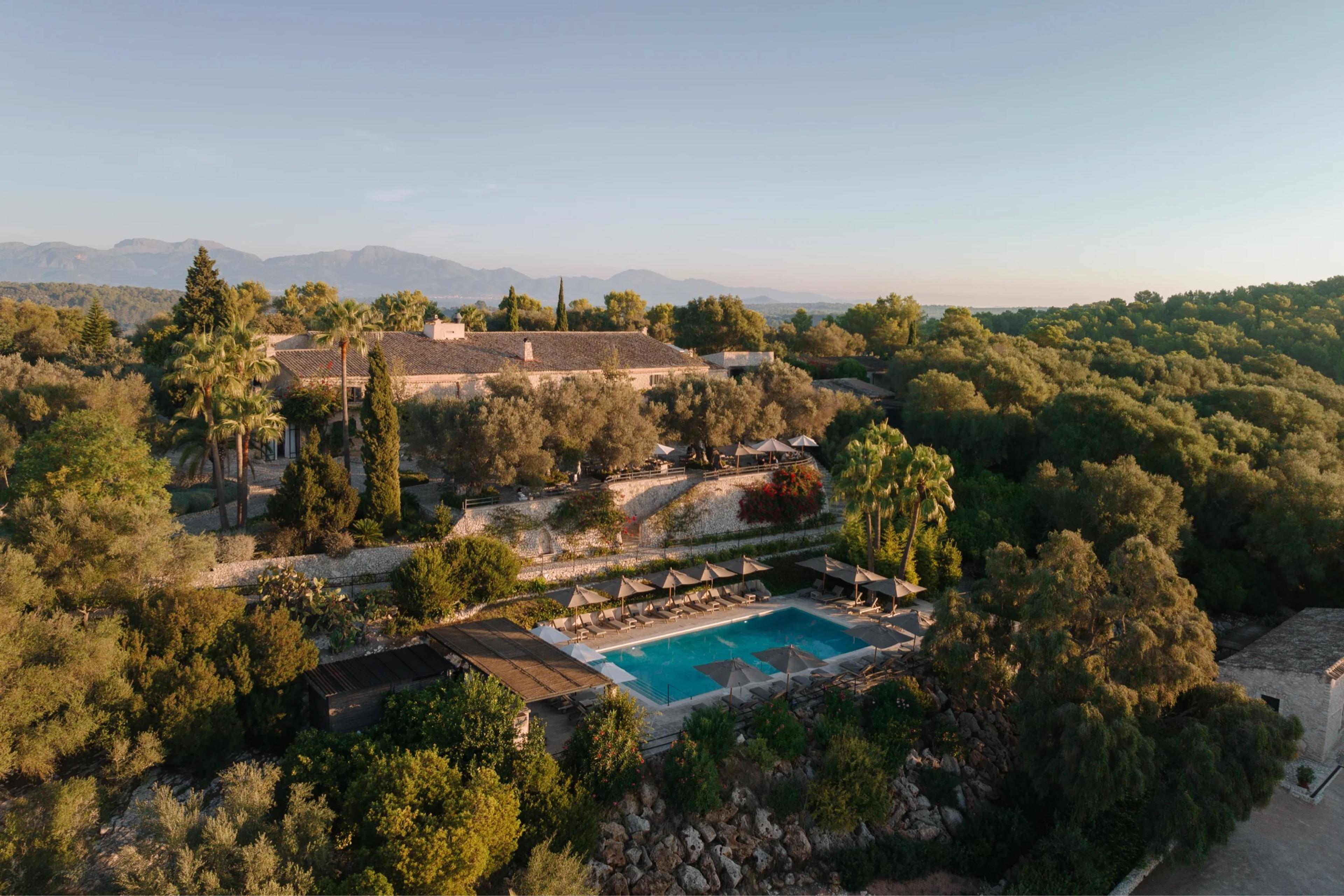 An aerial view of a stone hotel building, a swimming pool on a terraced garden, and trees, with mountains in the background.
