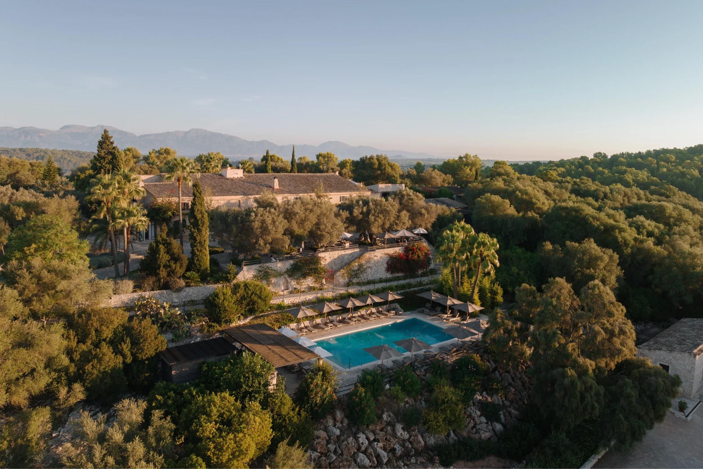 An aerial view of a stone hotel building, a swimming pool on a terraced garden, and trees, with mountains in the background.
