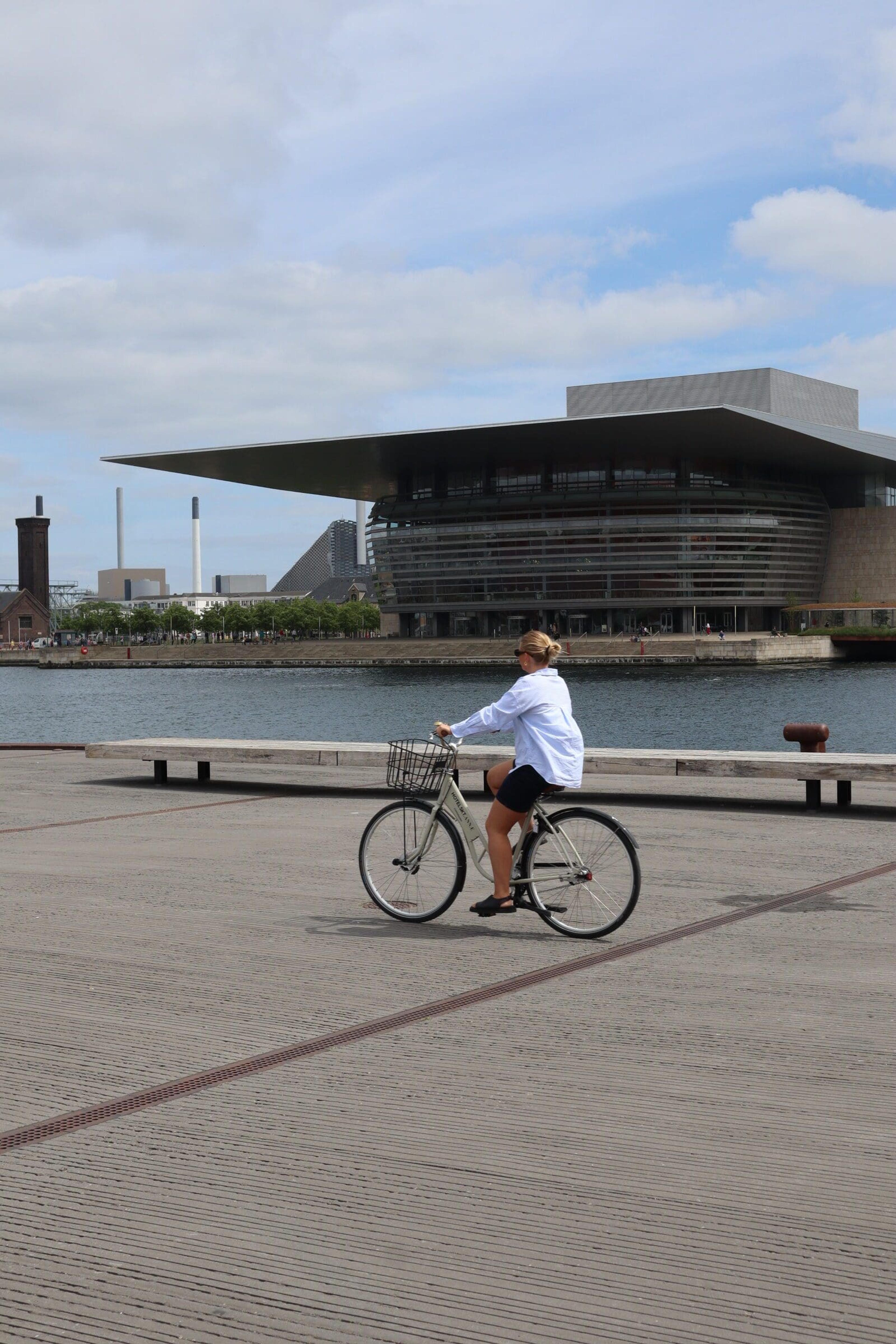 A woman rides a bicycle along a waterfront promenade with the Copenhagen Opera House across the water behind her.