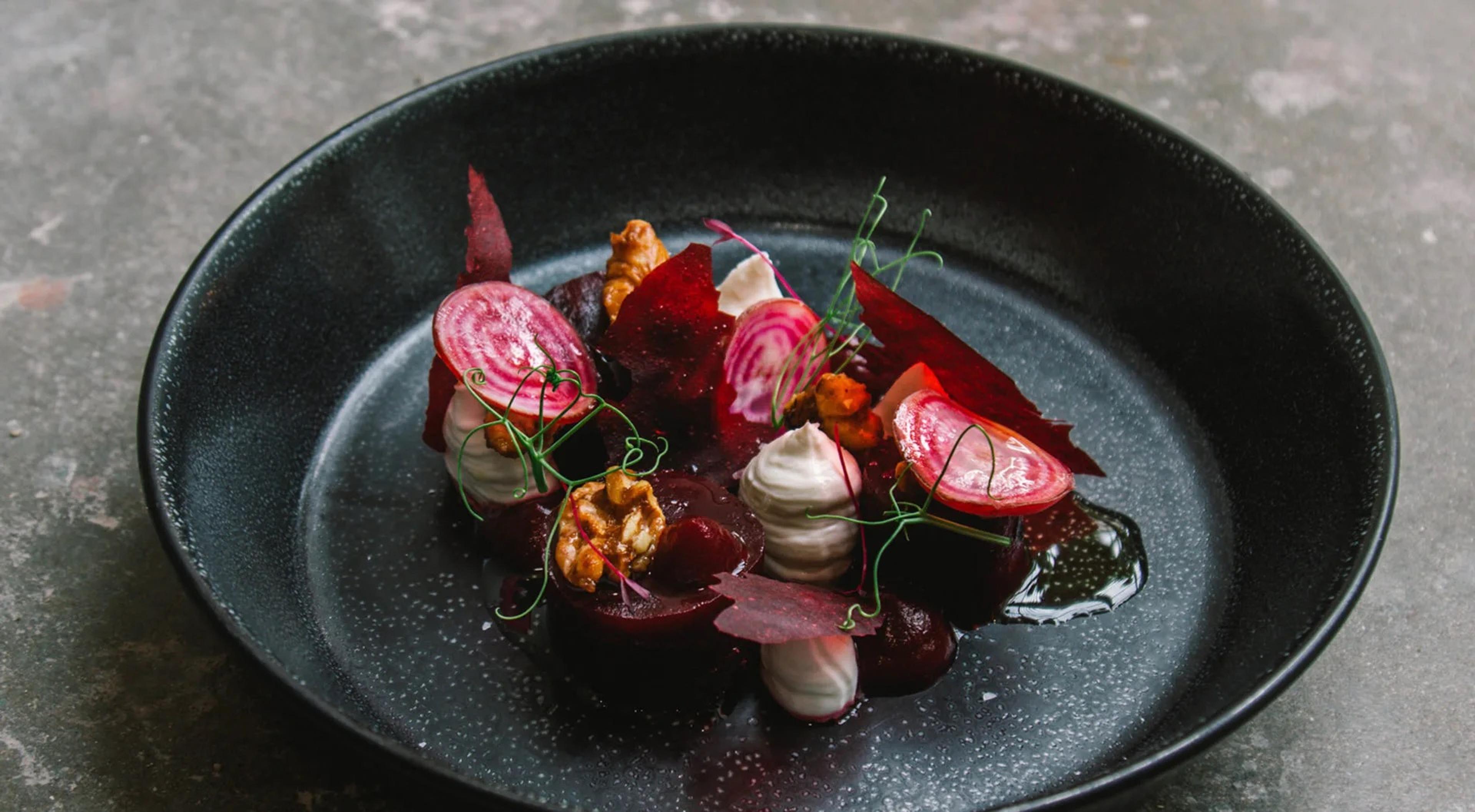 Beetroot and radish slices, walnut pieces, pale quenelles and cress on a dark plate, with a dark sauce pooled beside them.
