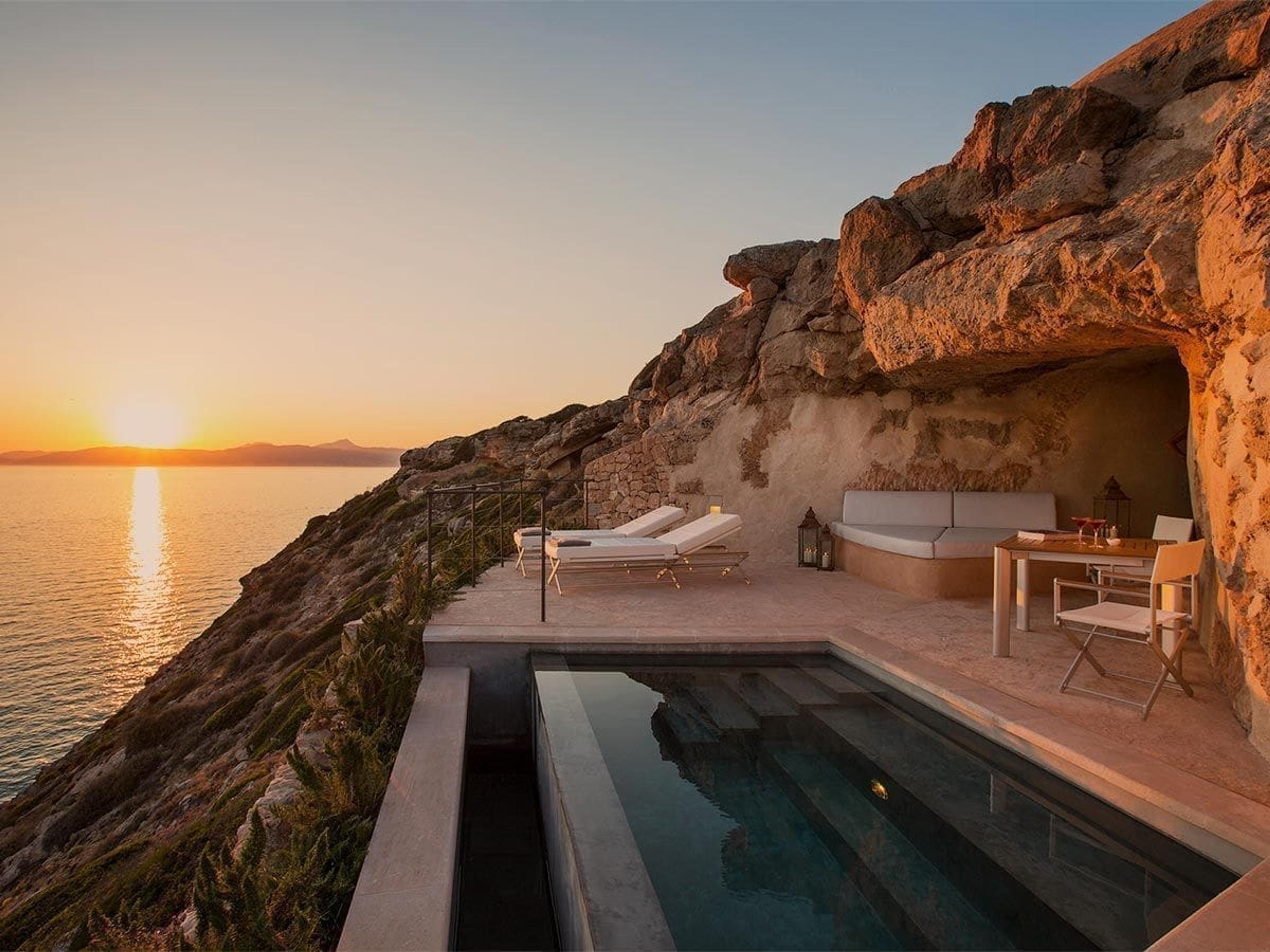 A plunge pool and terrace with sun loungers and a dining table built into a rock outcrop above the sea, lit by a setting sun.