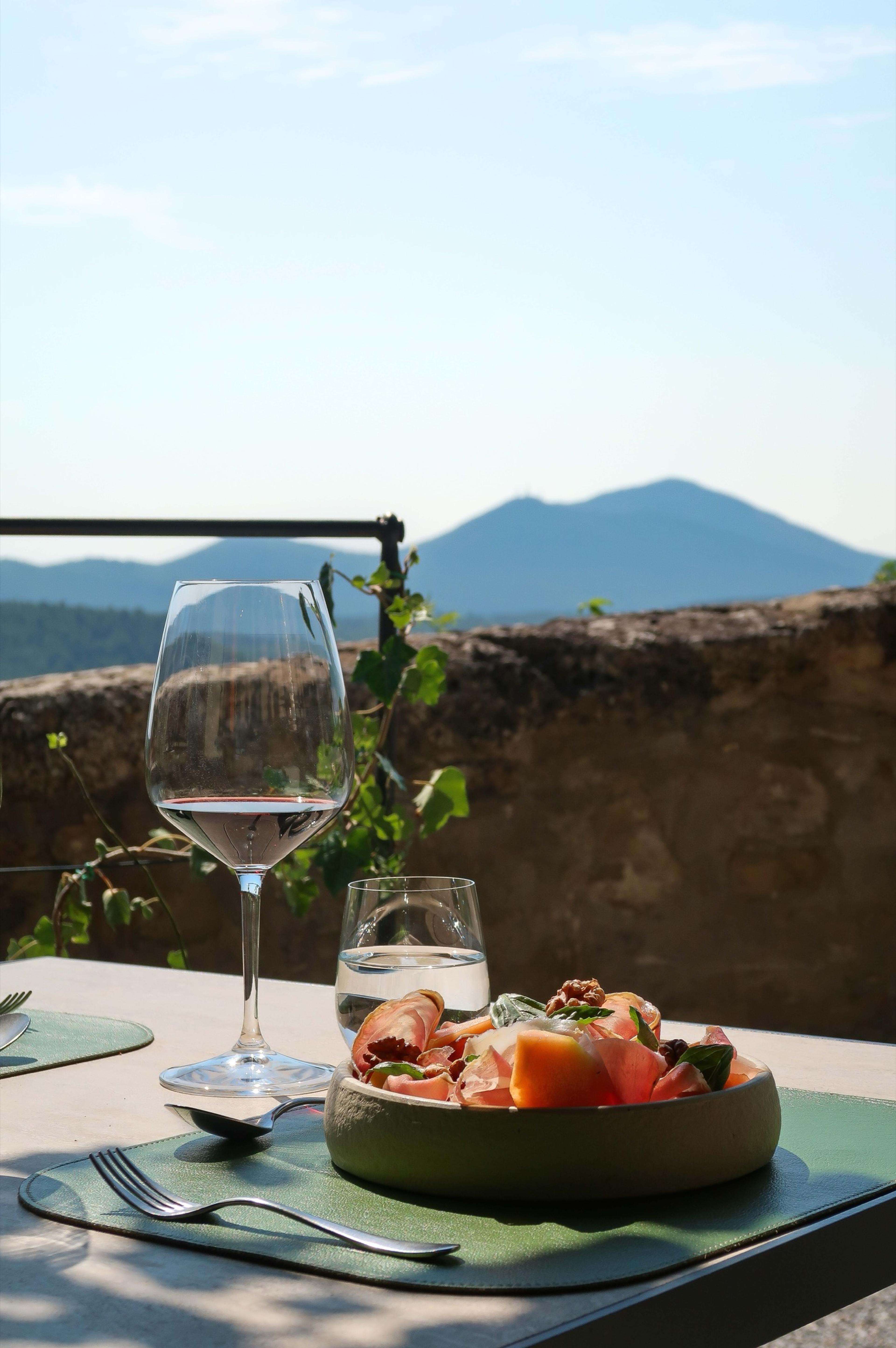 A table set with a glass of red wine, a glass of water, cutlery and a bowl of cured meat and melon, hills visible beyond a stone wall.