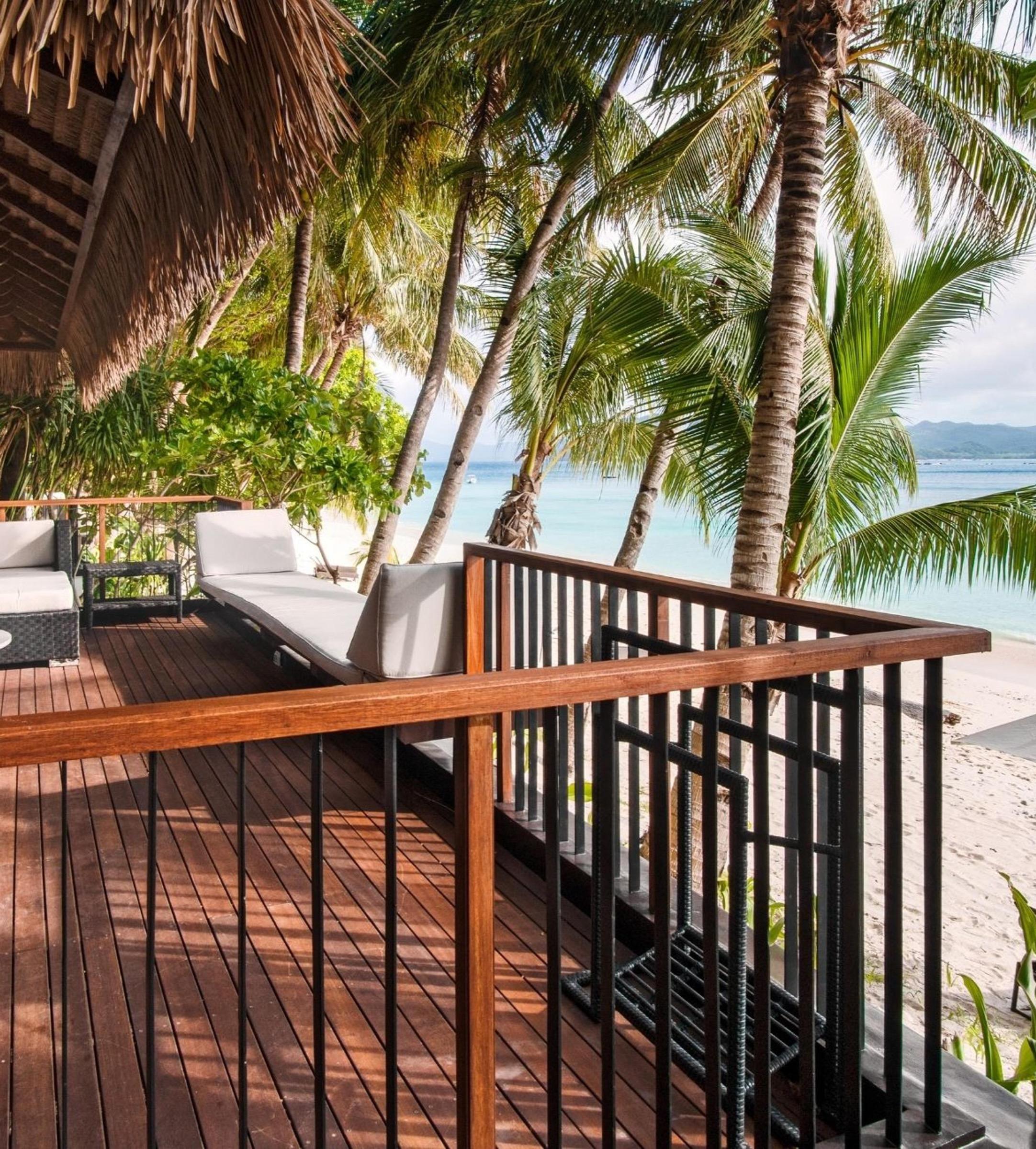 A wooden deck with cushioned loungers under palms, looking over the sand to the sea.