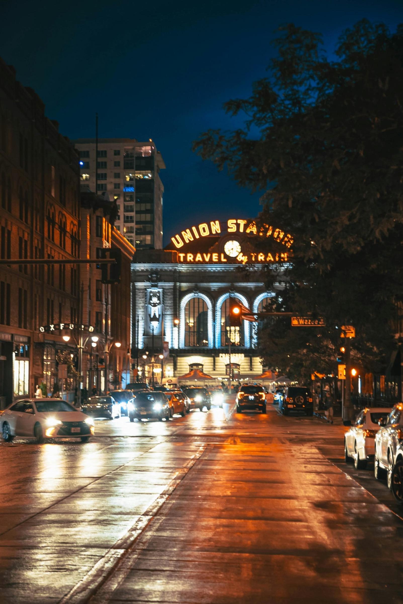 A city street at night in the rain, with traffic and the lit sign of Union Station above an arched entrance.