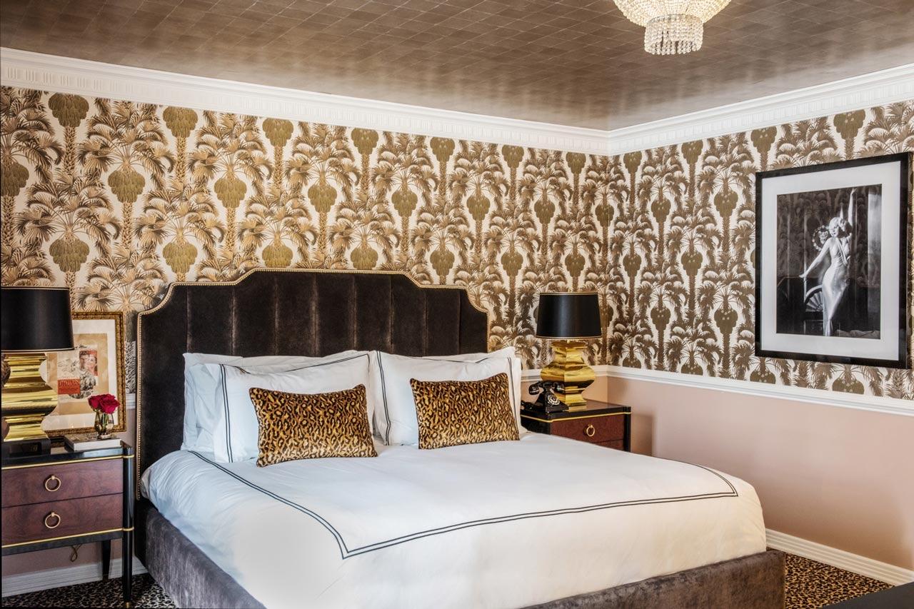 A bedroom with a tufted dark brown headboard, a white bed dressed with a leopard print cushion, gold palm wallpaper, and a tiled gold ceiling with a small chandelier.