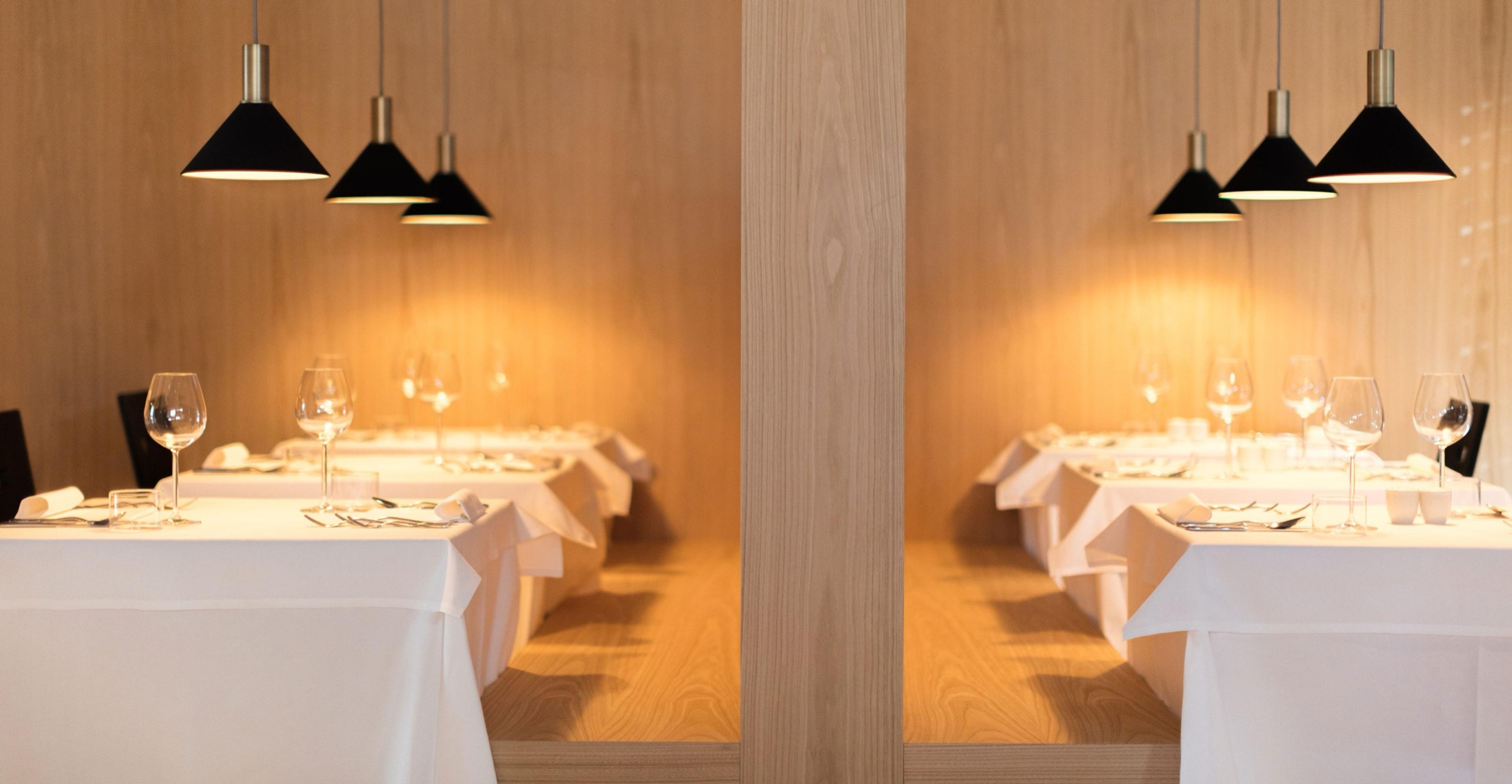 A hotel restaurant with two mirrored rows of white-clothed tables set with wine glasses, under black pendant lamps against wood panelling.