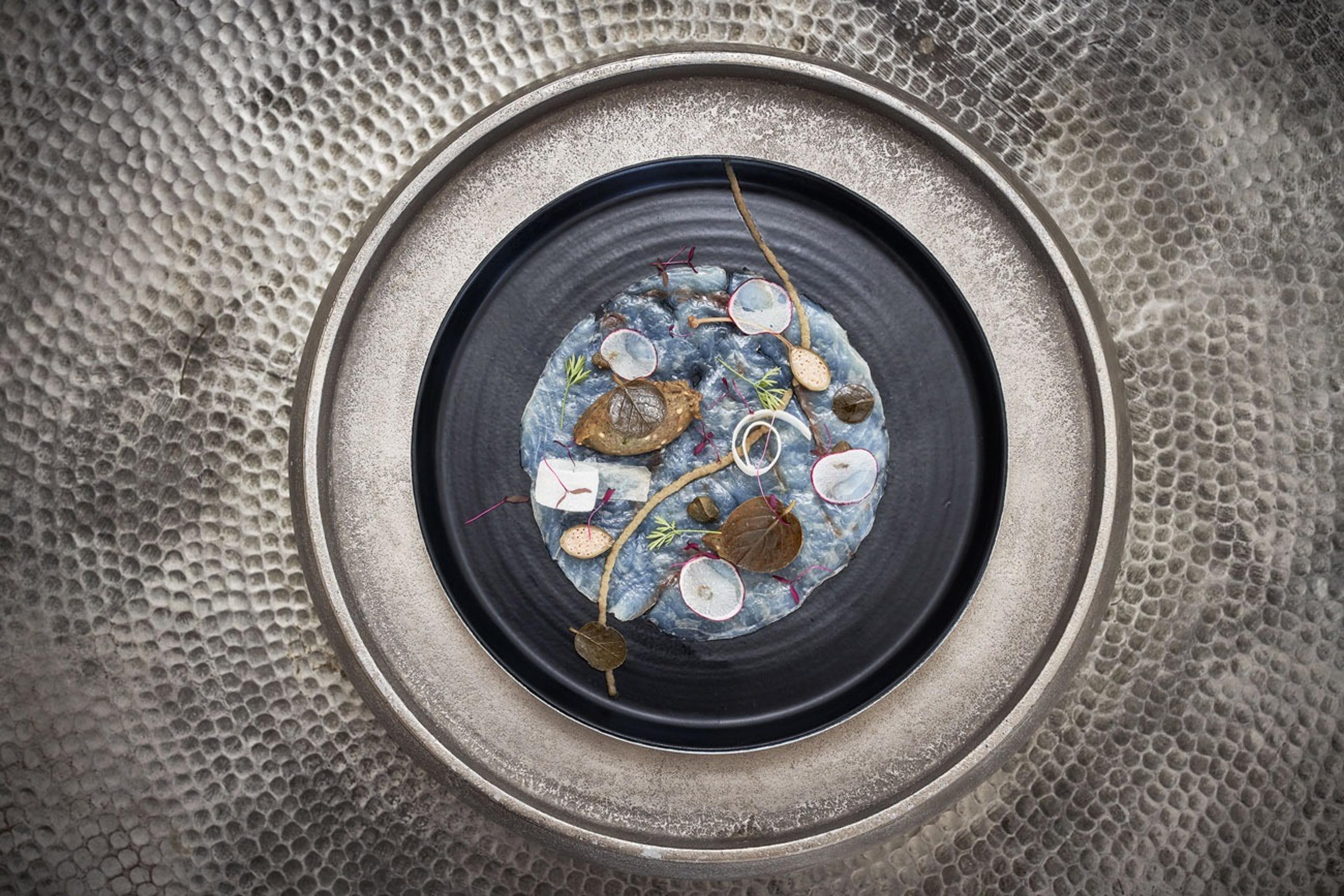 Overhead, a blue-grey dish with radish slices, herbs and a crisp leaf, set on a dark metal plate.