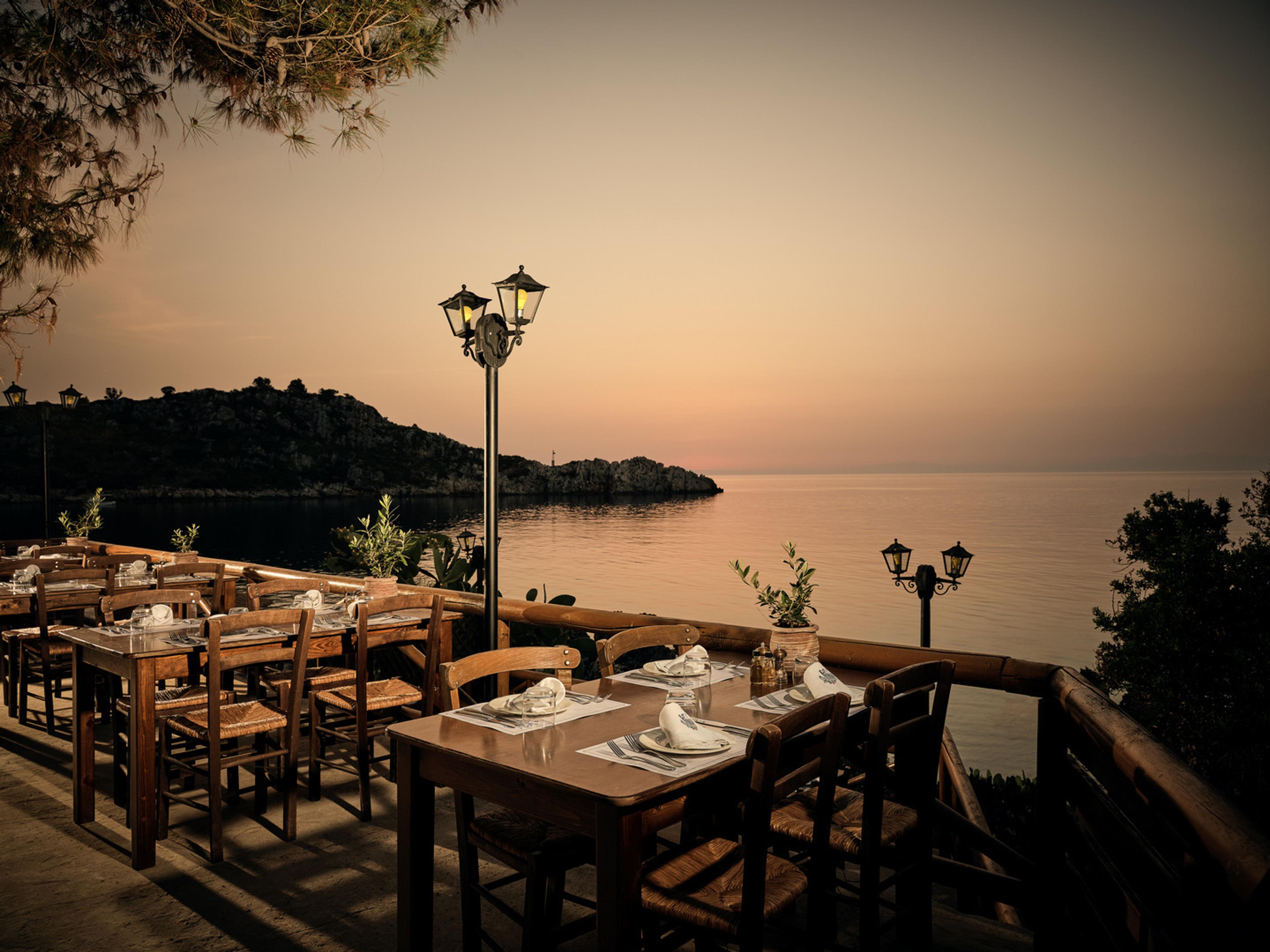 Tables laid for dinner on a stone terrace above the sea at dusk, lamp posts along the railing and a wooded islet offshore