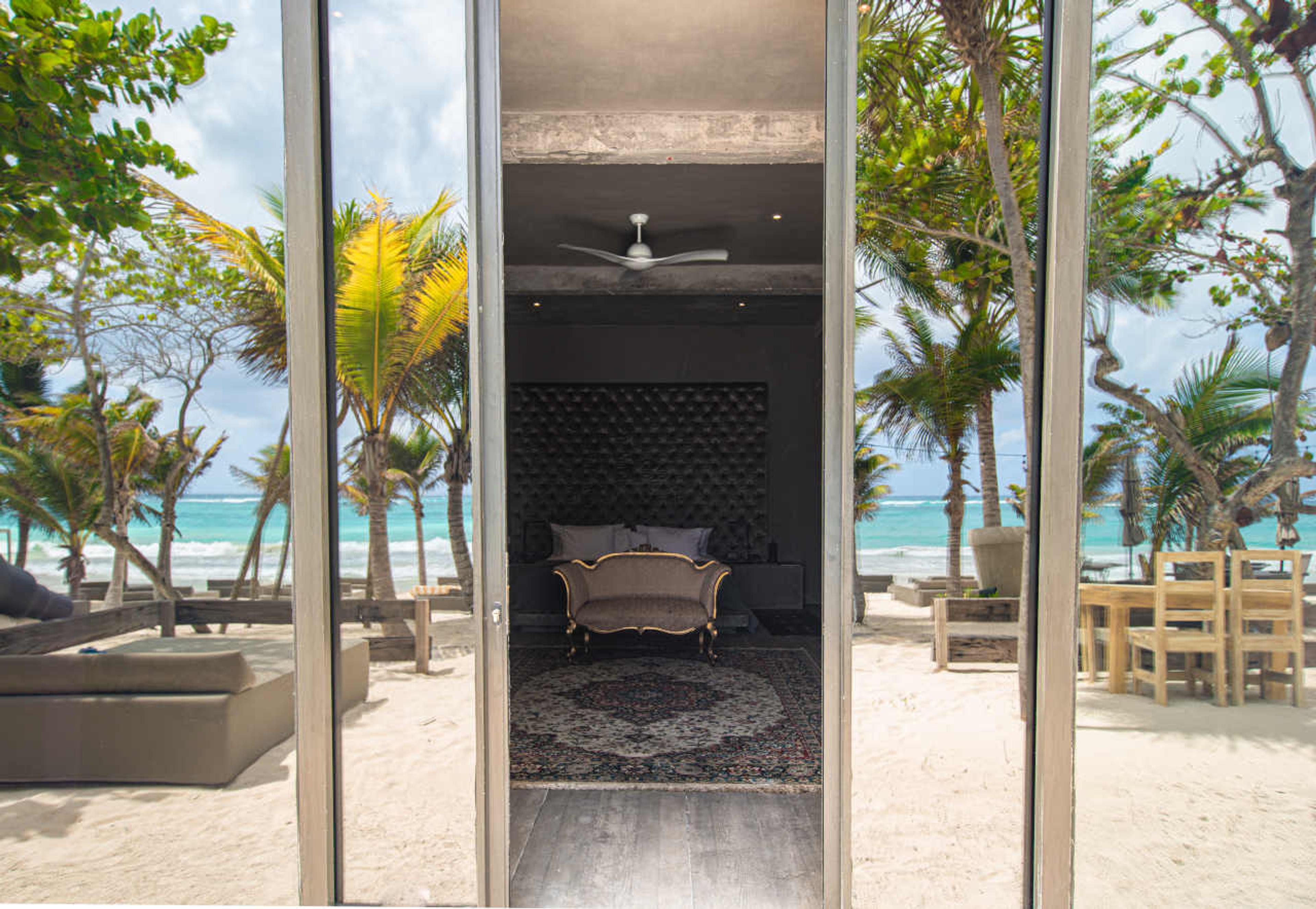 A view through glass doors from a beach terrace into a bedroom with a dark tufted headboard and a small sofa.